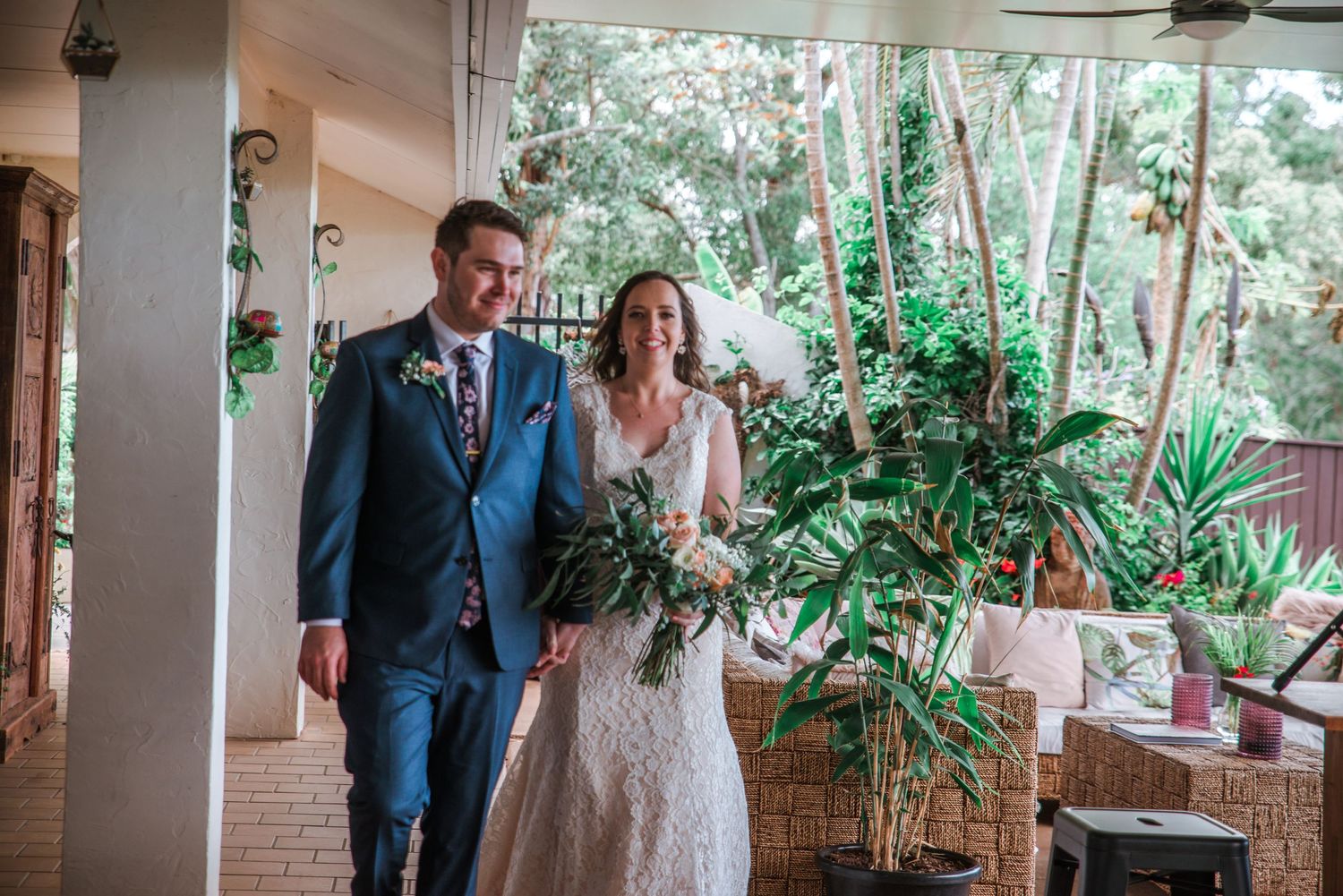 Wedding couple walking through covered porch surrounded by potted plants.