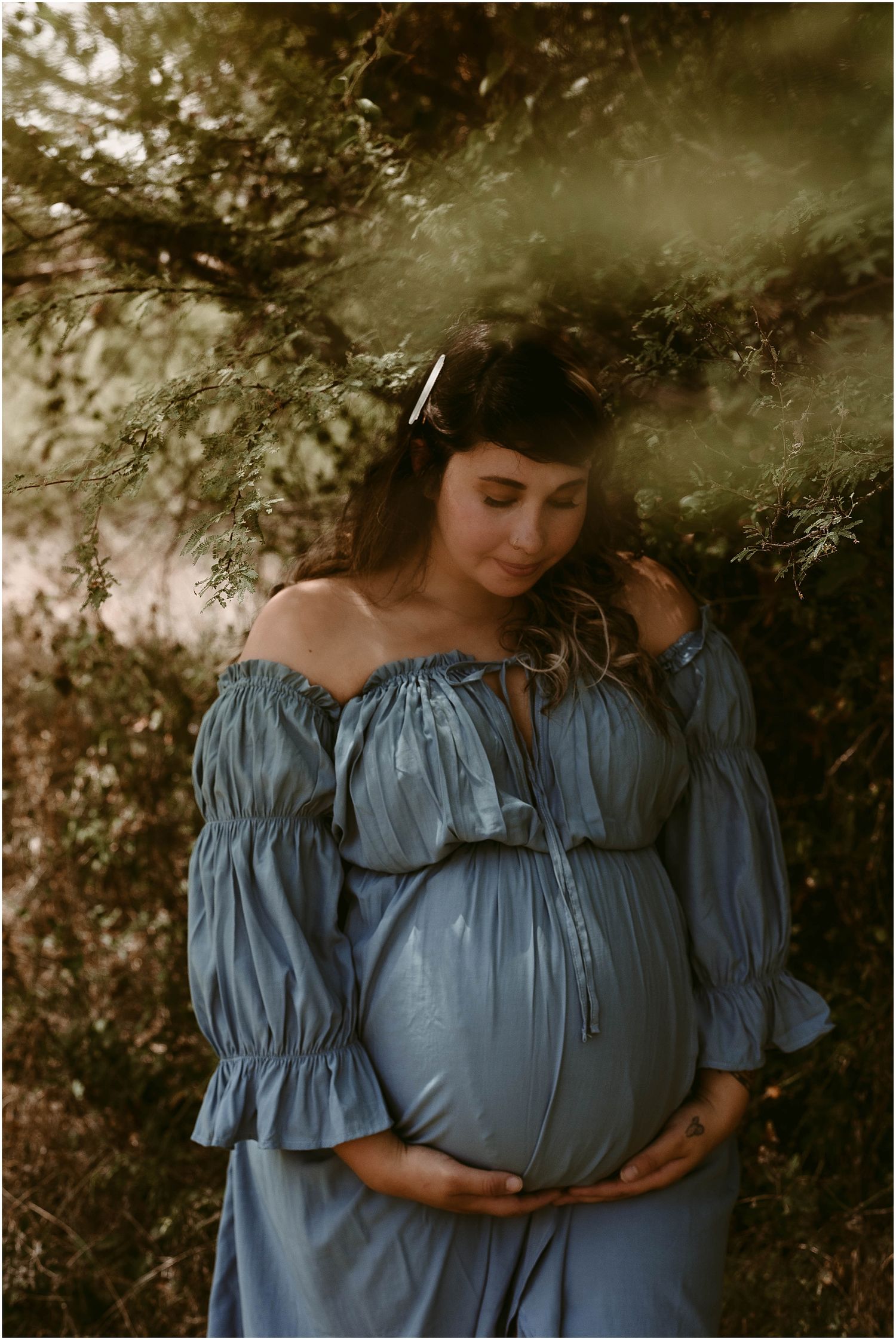 Dreamy maternity photo with dappled lighting through trees and blue peasant dress.