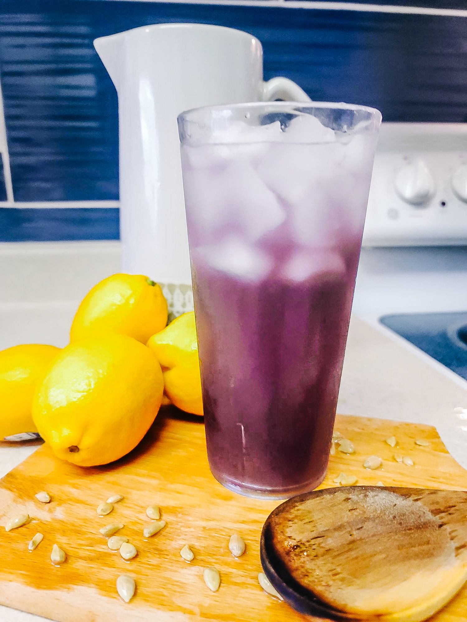 A purple beverage with ice sits next to fresh lemons on a wooden cutting board.