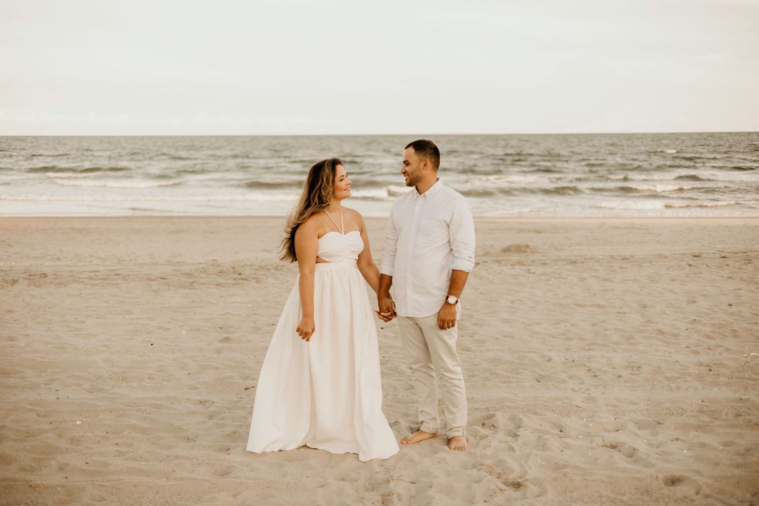 Couple in white attire standing together on sandy beach at golden hour.