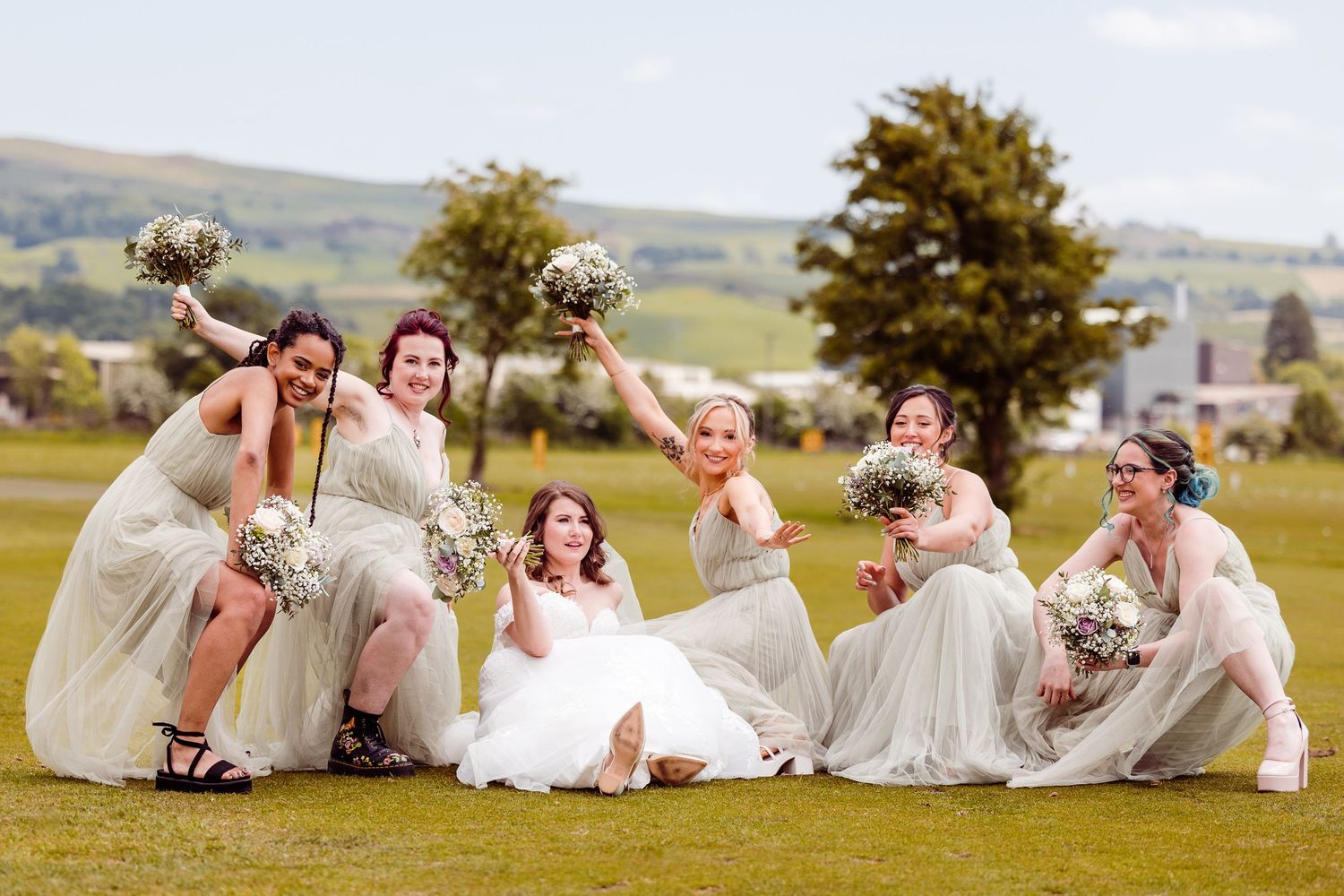 Wedding party in light dresses pose playfully on a grassy hillside with bouquets and raised arms.