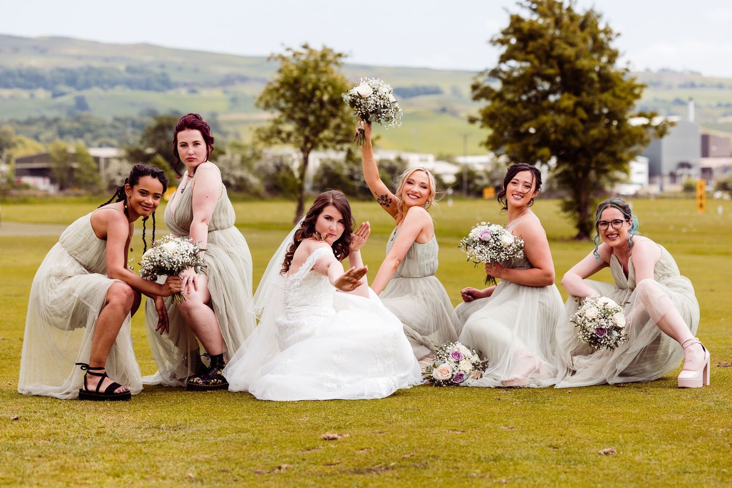 Bridal party sitting on grass with bouquets strike a fun celebratory pose together.
