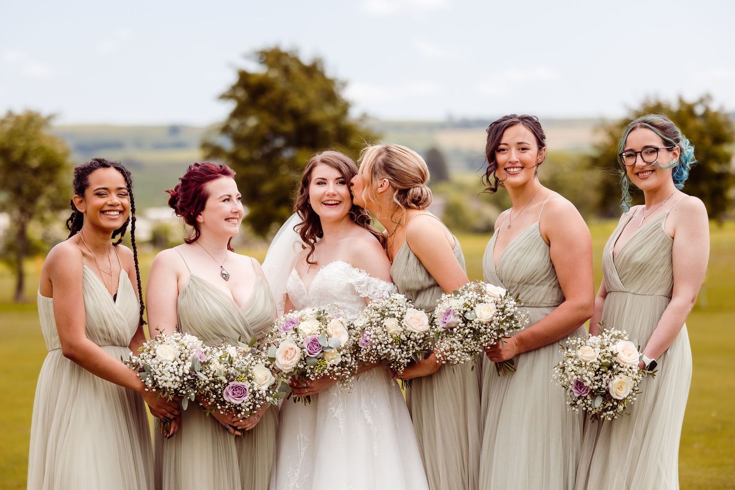 Bridesmaids in neutral colored dresses holding floral bouquets pose on golf course.