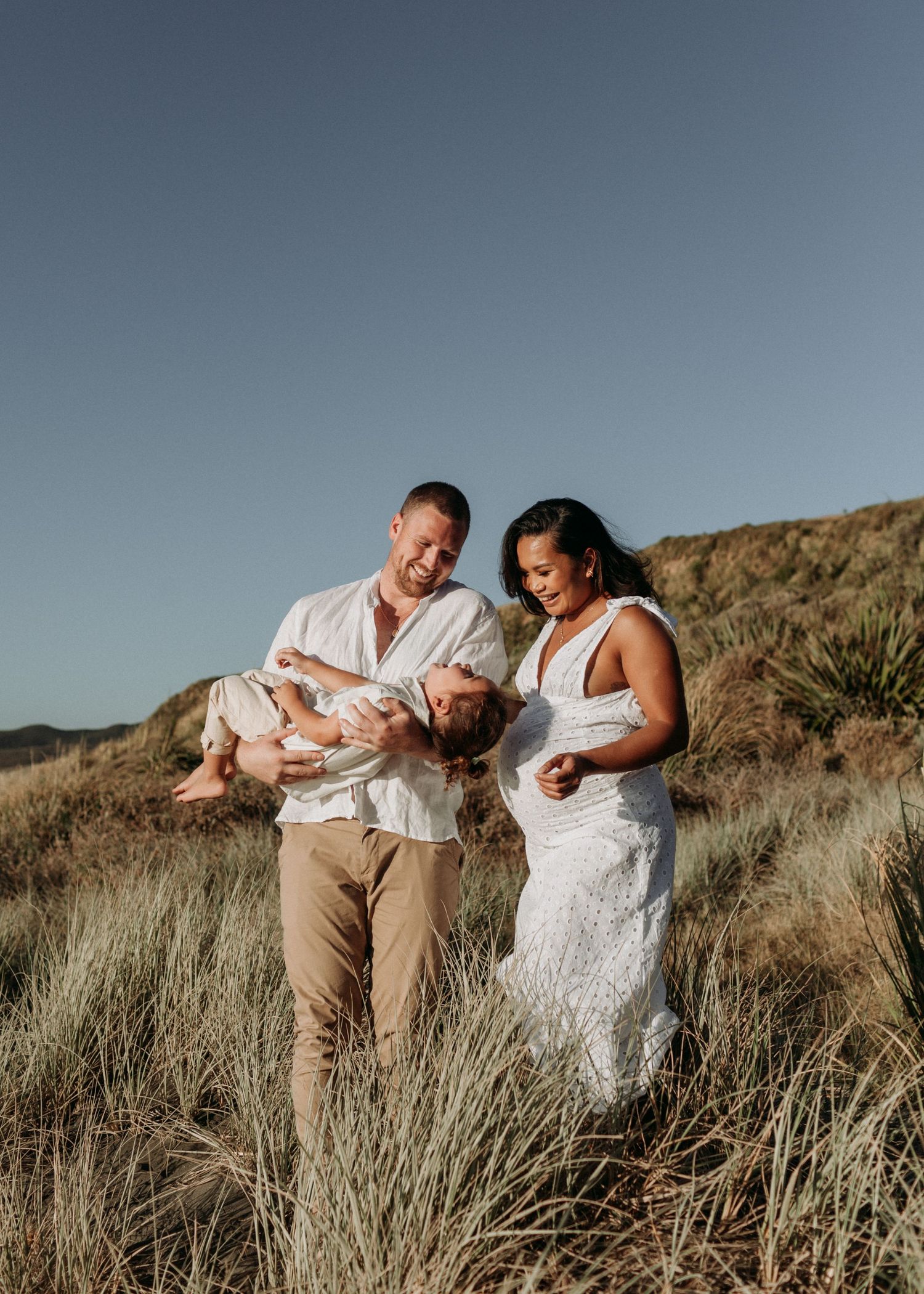 A couple in white outfits shares intimate moments among coastal grass and dunes.