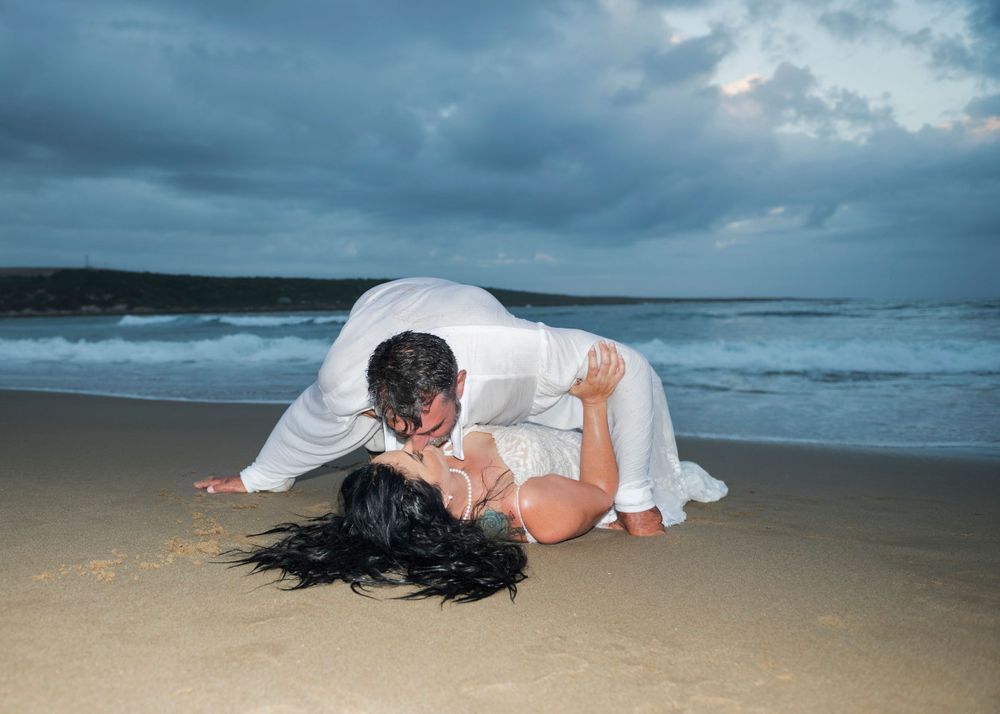 A romantic couple in white sharing an intimate moment on a beach with dramatic stormy skies overhead.