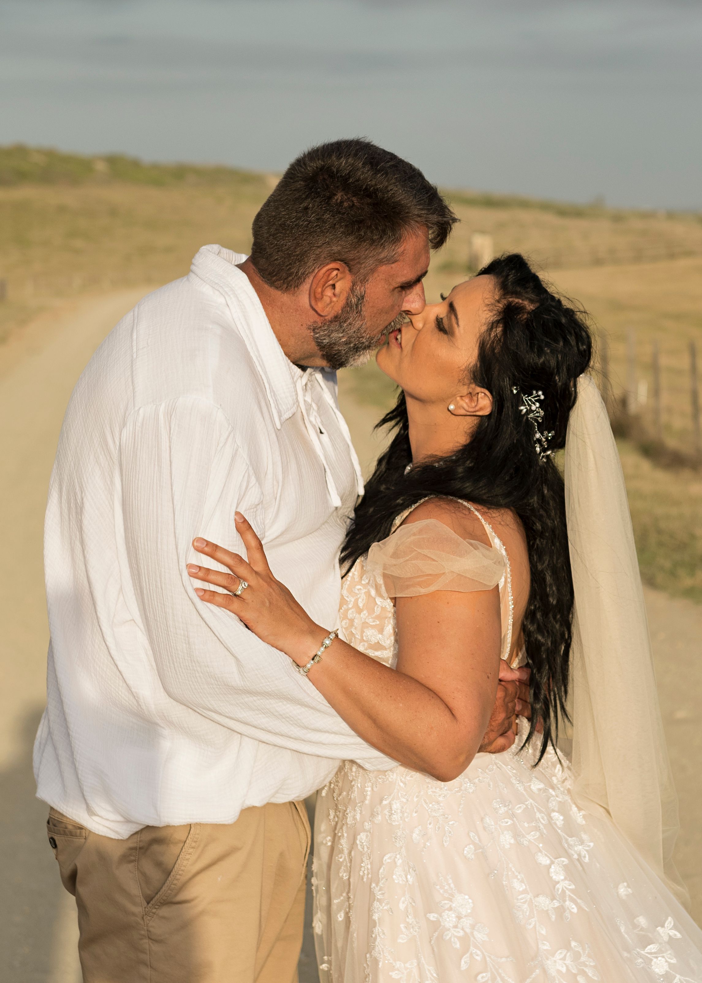 A wedding couple sharing a loving kiss outdoors in formal white attire at golden hour.