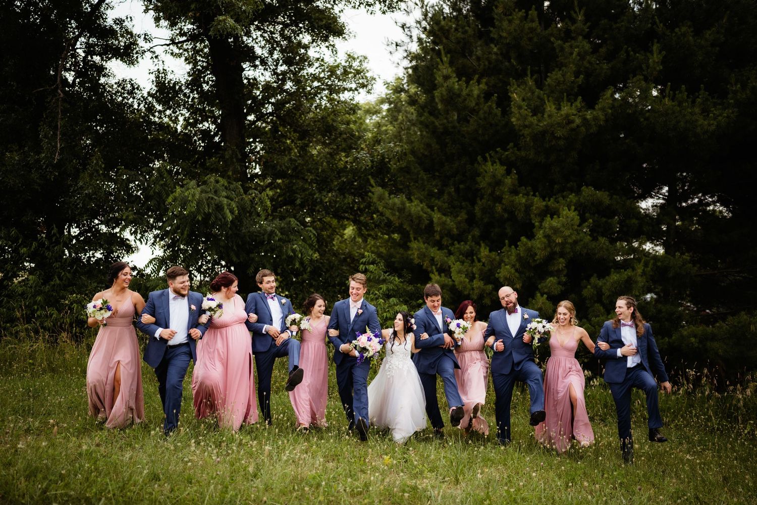 Wedding party in pink dresses and navy suits pose together in a line on grassy meadow with trees in background.