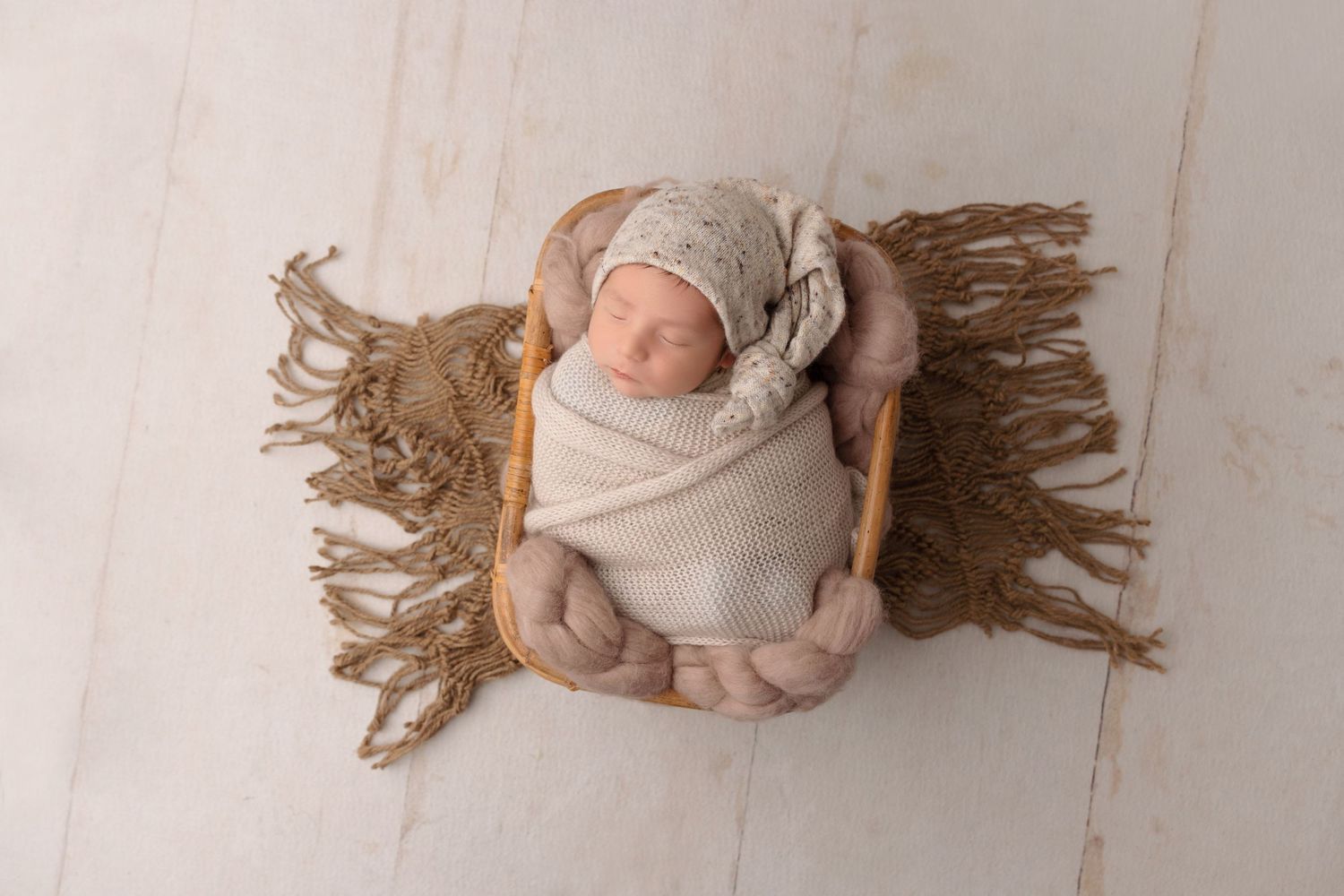 Newborn wrapped in beige cloth lying on textured wooden surface.