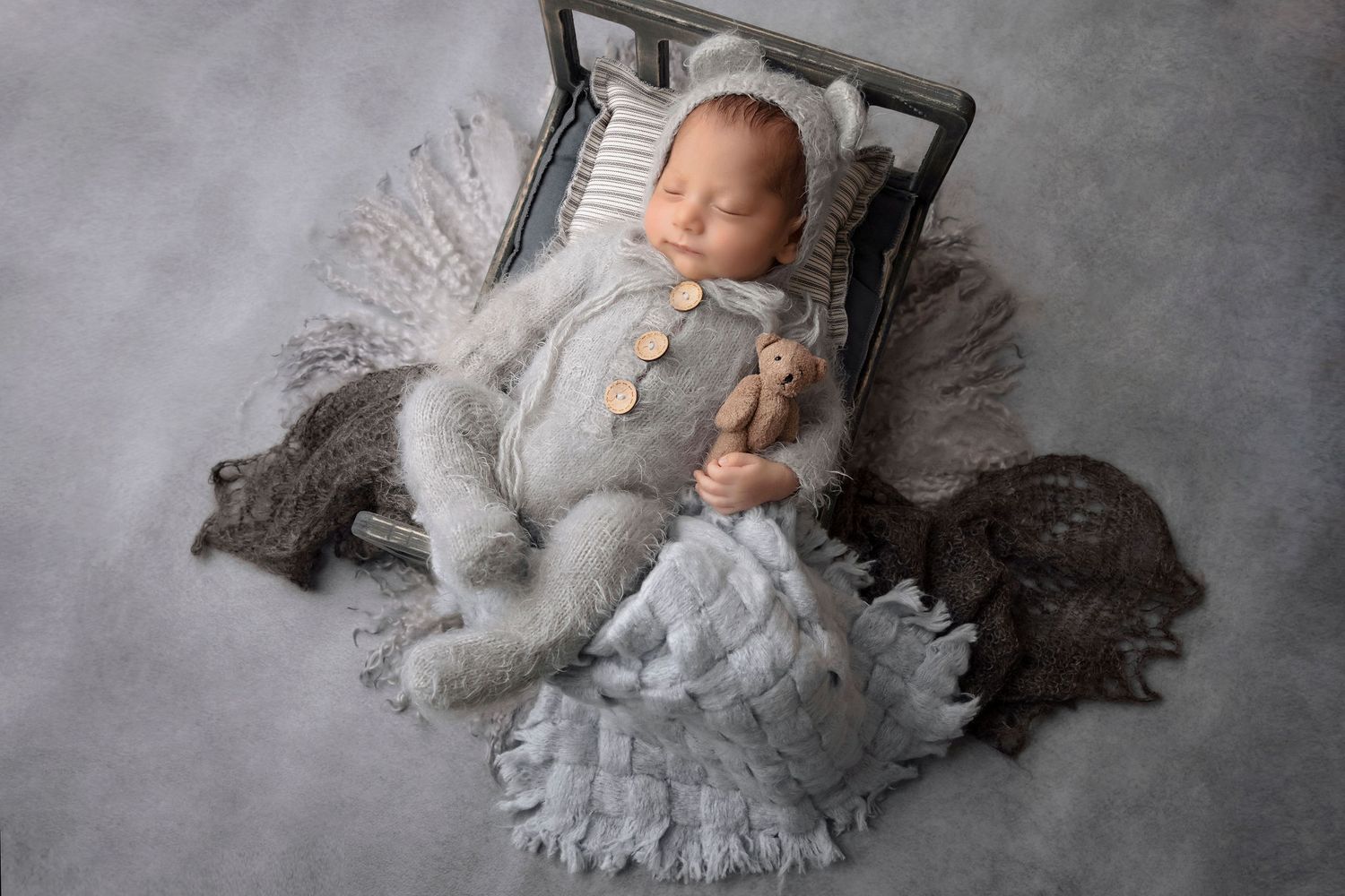 Sleeping newborn in gray romper posed on textured layers in vintage-style bed prop.