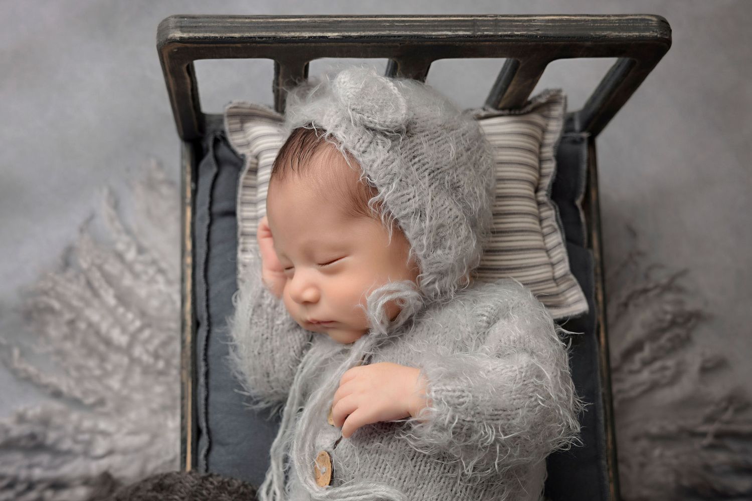 Sleeping newborn wearing soft gray bonnet posed in rustic wooden bed prop.