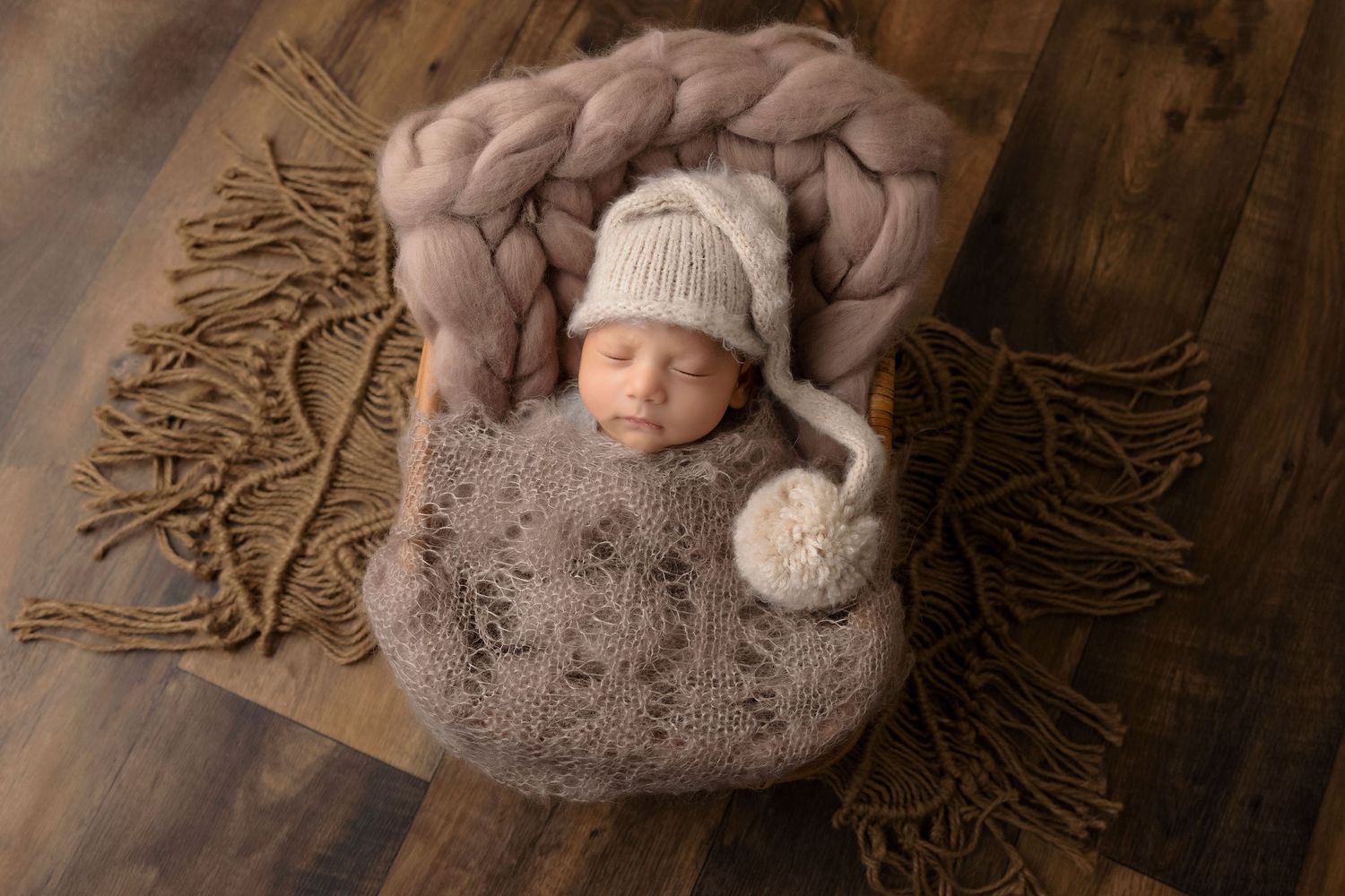 Sleeping newborn in knit sweater and hat posed in cozy basket on wooden floor.
