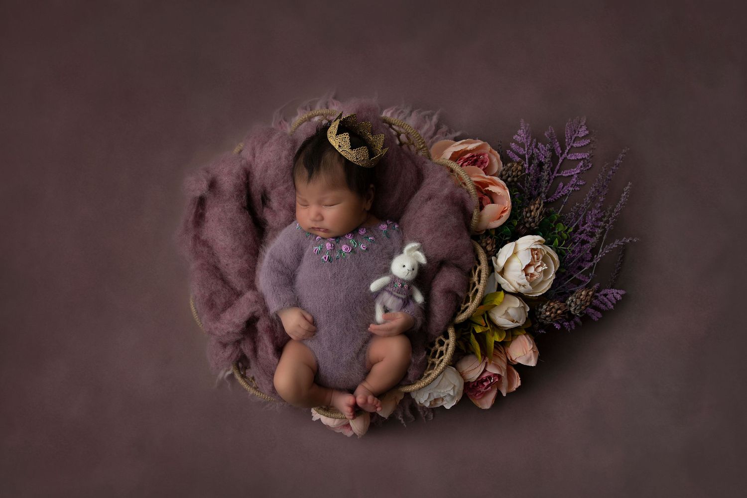 Newborn photography featuring a sleeping baby nestled in purple fabric with floral decorations.