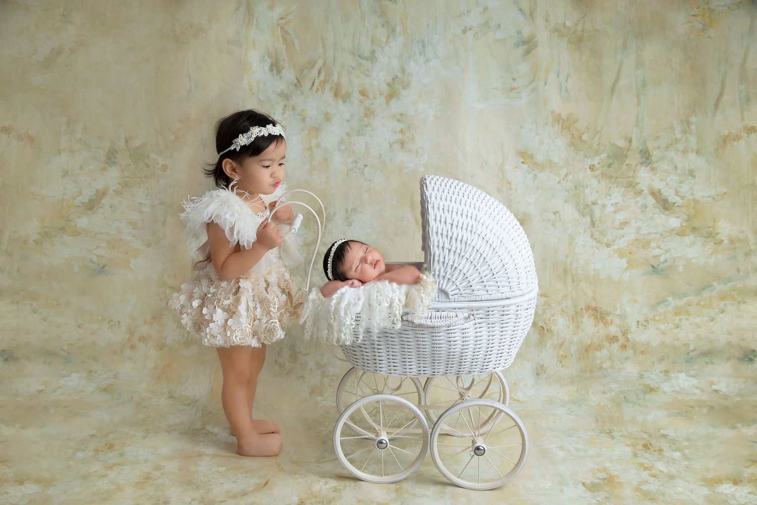 Children's portrait with vintage white pram and toddler in lace dress against textured wall.