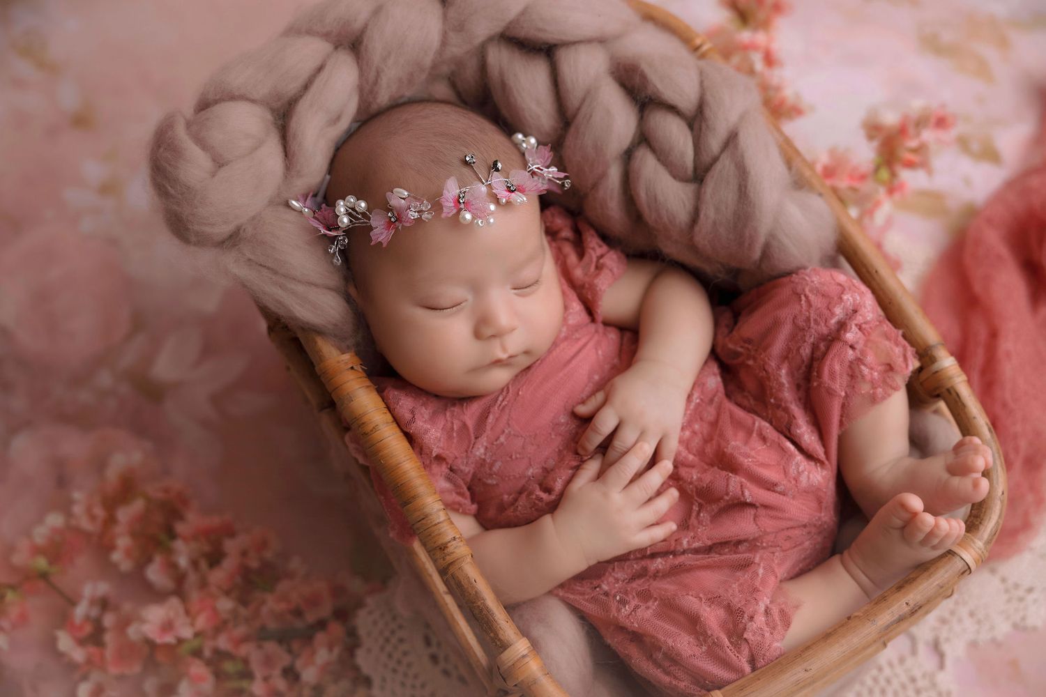 Sleeping newborn in pink outfit with floral crown resting in wooden prop bed.