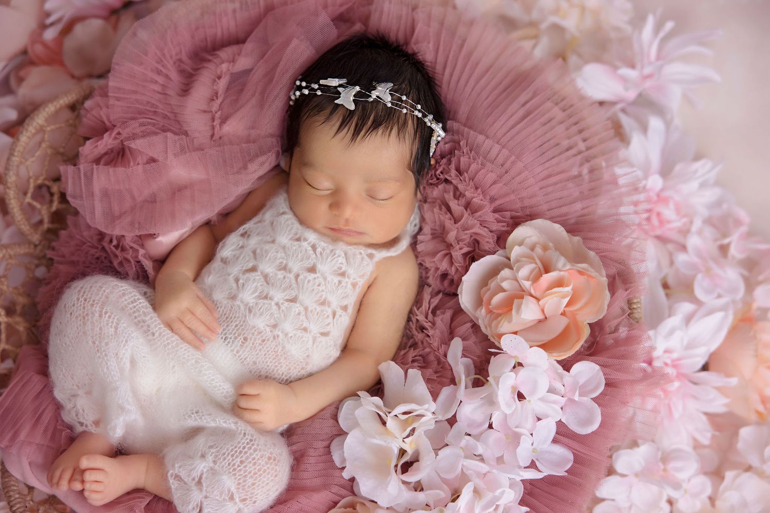 Peaceful newborn resting on pink lace surrounded by soft pink flowers and wearing dainty headpiece.
