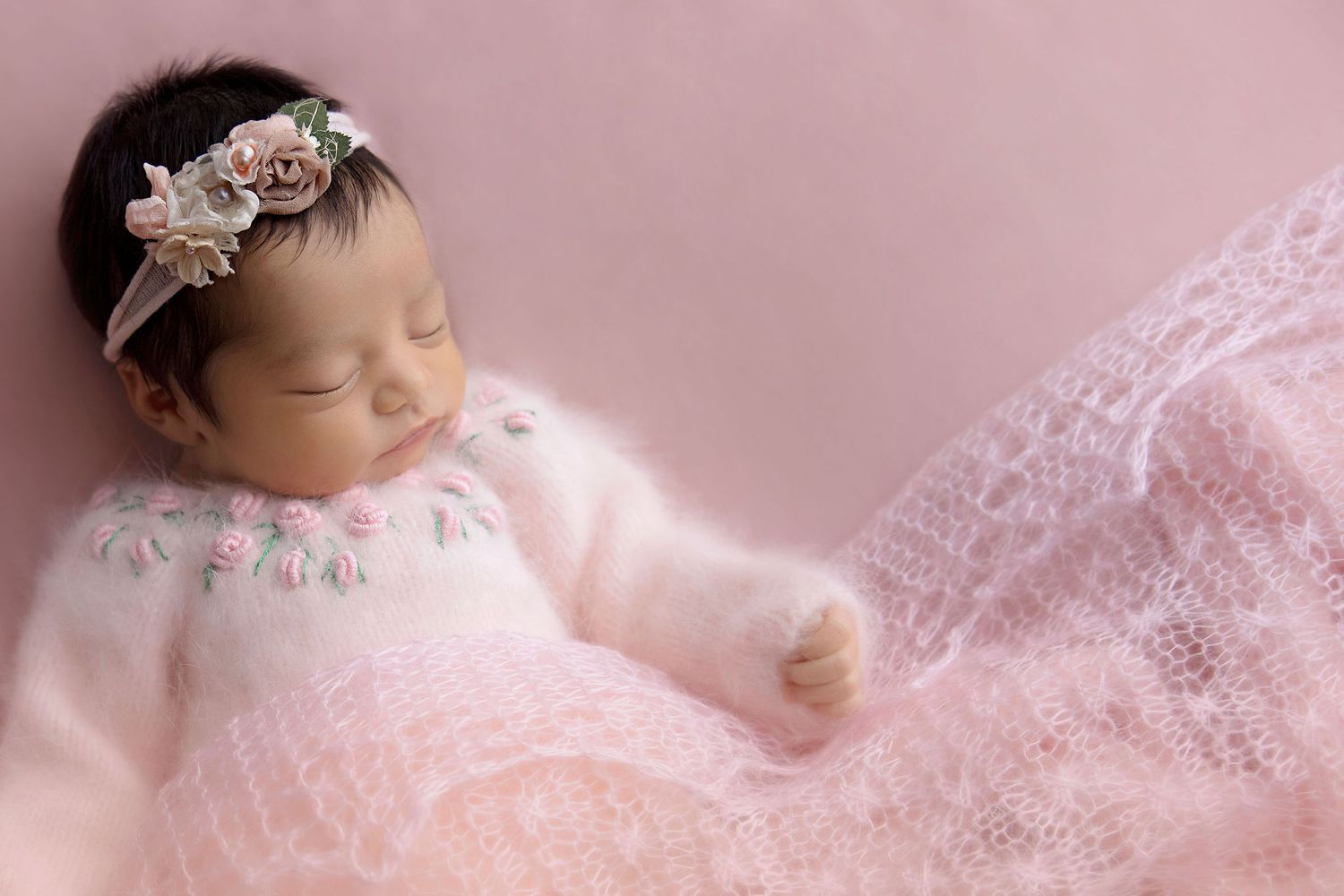 Sleeping newborn in pink lace wrap wearing delicate floral headband against soft pink background.