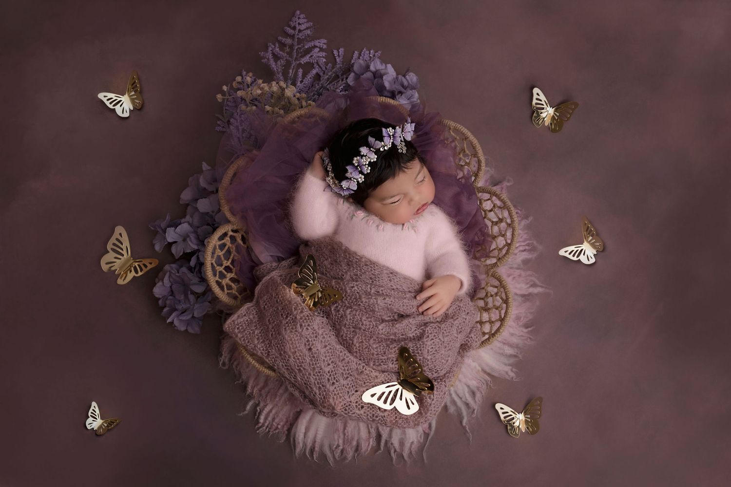 Artistic overhead shot of sleeping newborn surrounded by butterflies on purple backdrop.