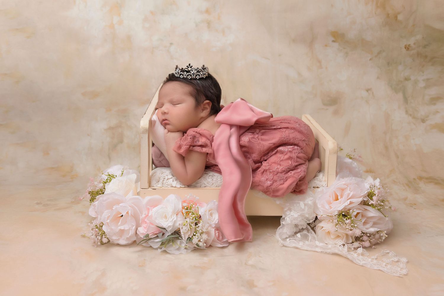 Sleeping baby in pink dress and crown posed with delicate flowers on beige backdrop.