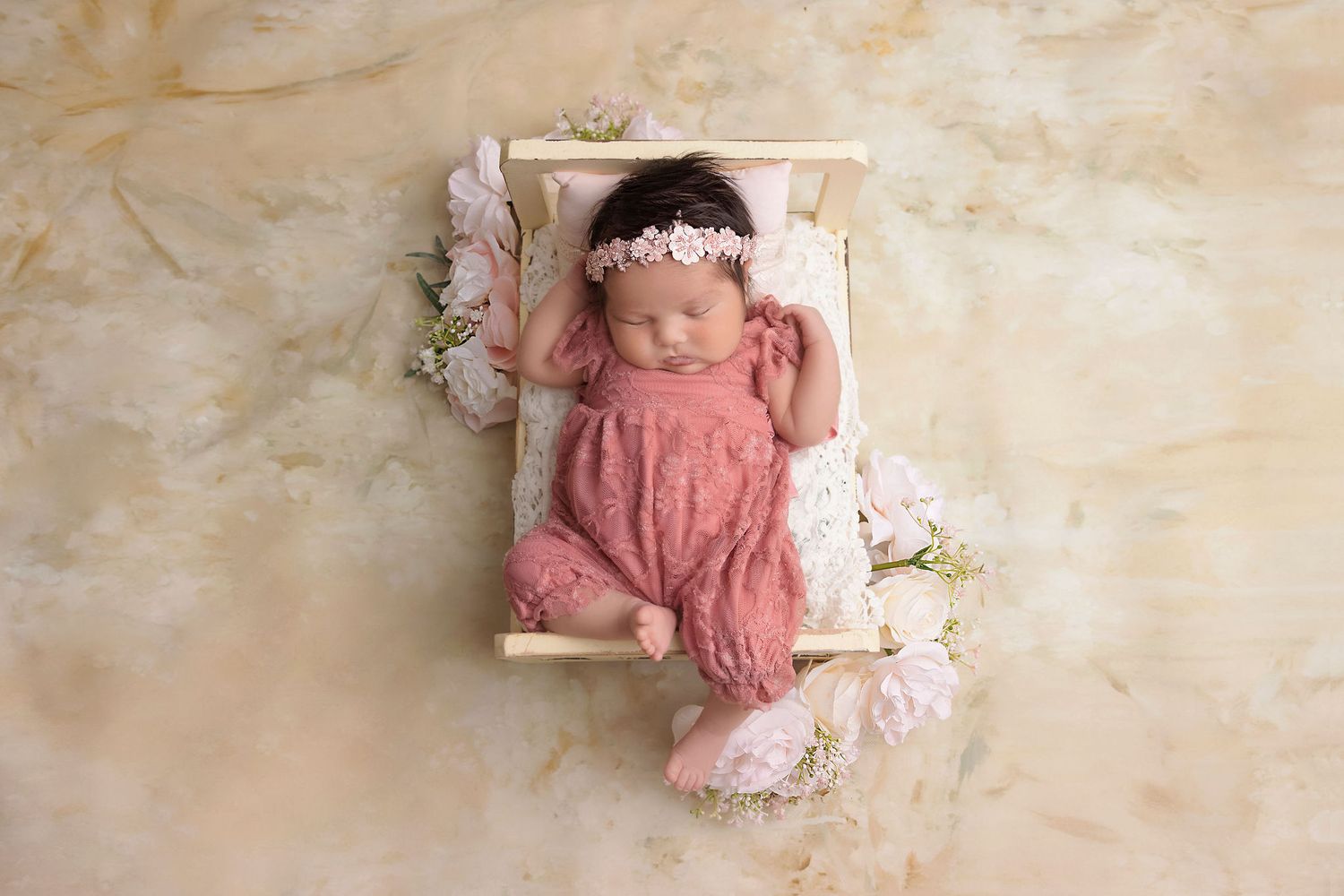 Newborn sleeping in pink romper on miniature bed with floral accents.