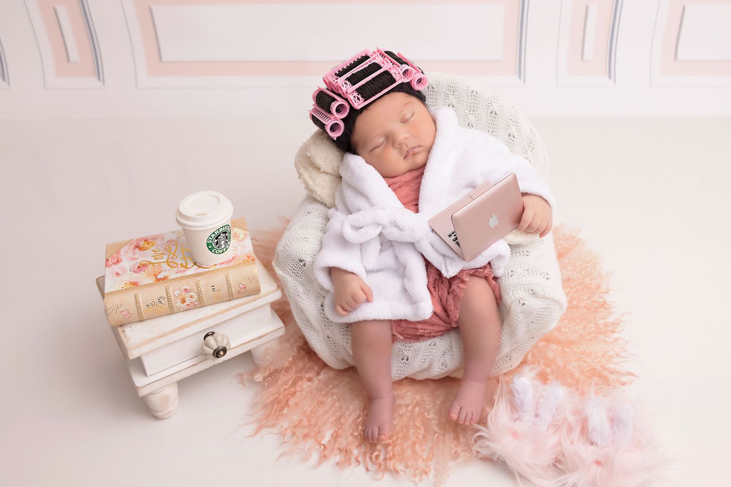 Newborn dressed in white robe resting on peach-colored blanket with wooden blocks and decorative accessories.
