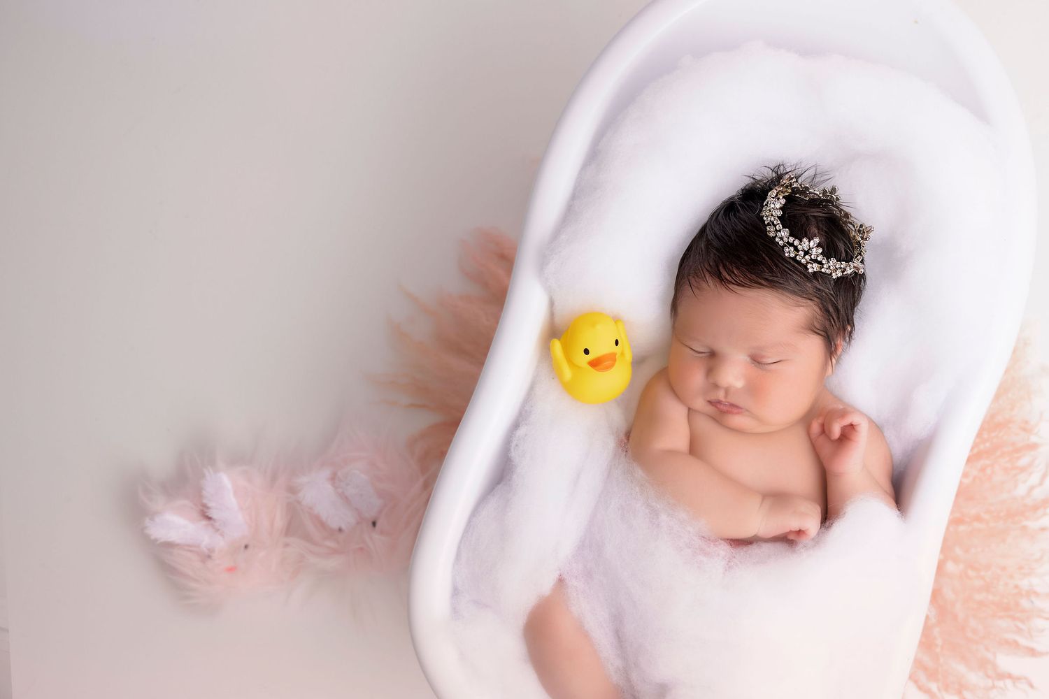 Sleeping newborn in white bathtub with rubber duck and delicate crown accessory.