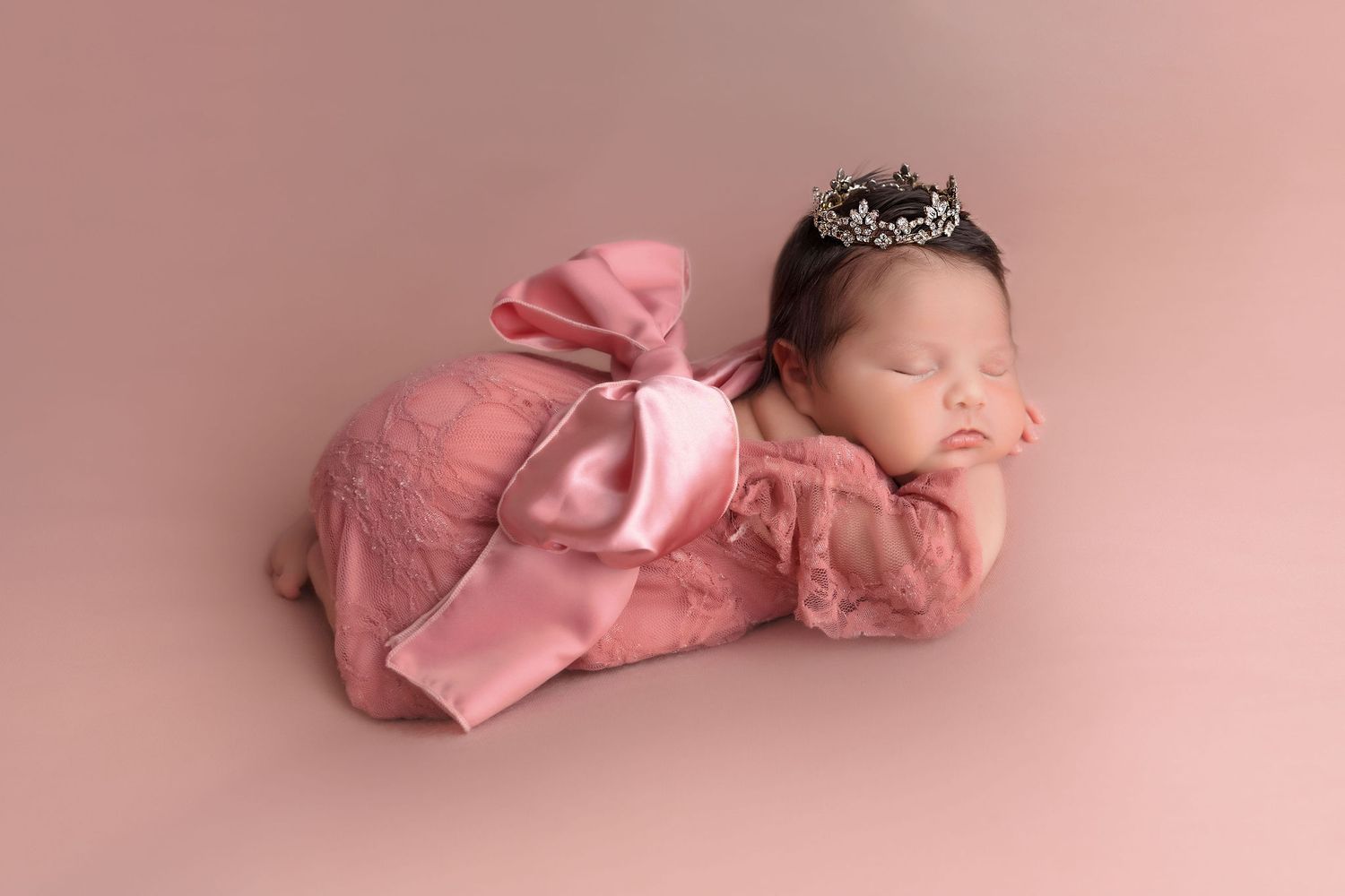 Sleeping newborn in pink lace dress with large bow and crystal tiara against rose background.