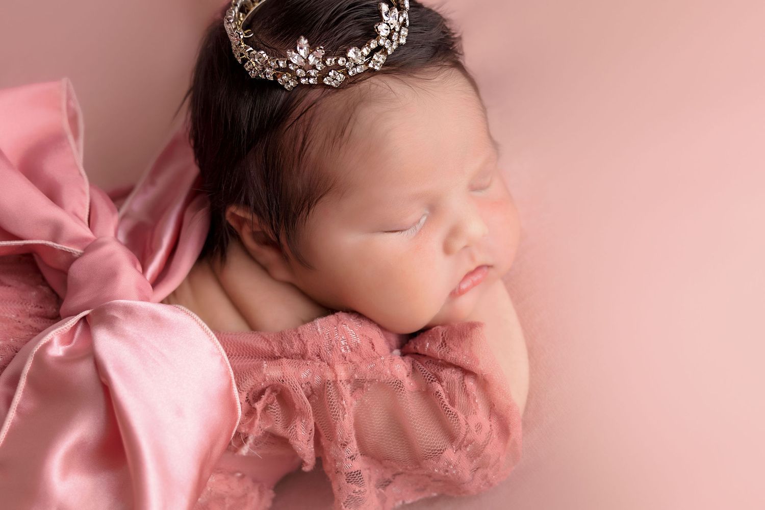 Peaceful newborn wearing tiara and pink dress with bow detail against rose background.