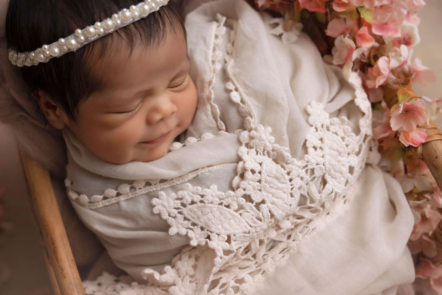 Sleeping newborn in white lace dress with pearl headband and floral decor.