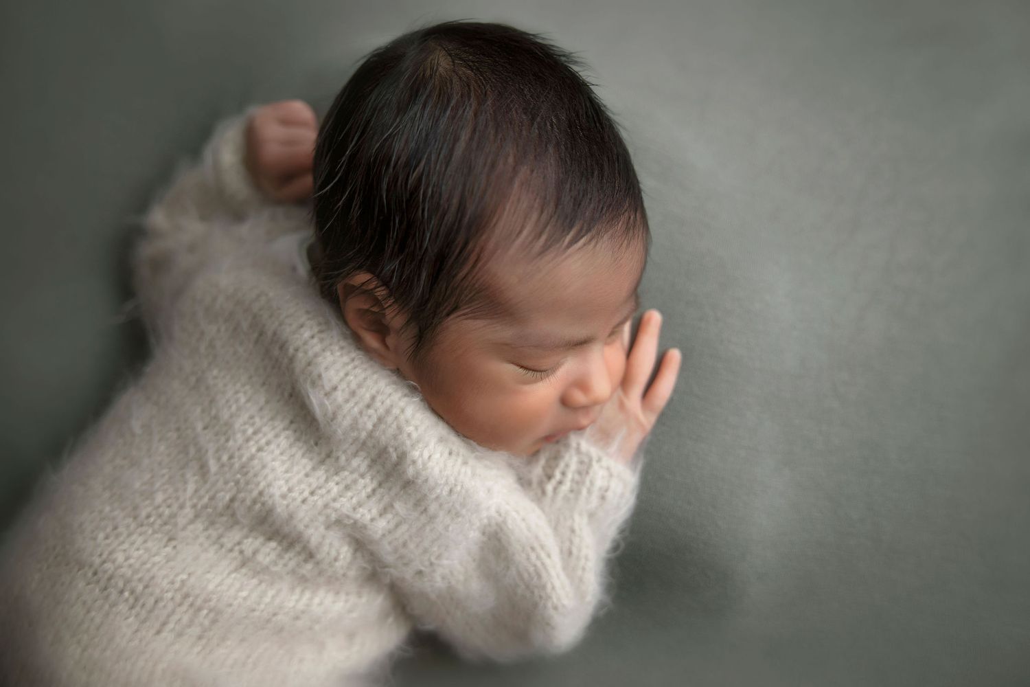 Peaceful newborn wrapped in soft gray knit sweater against neutral background.