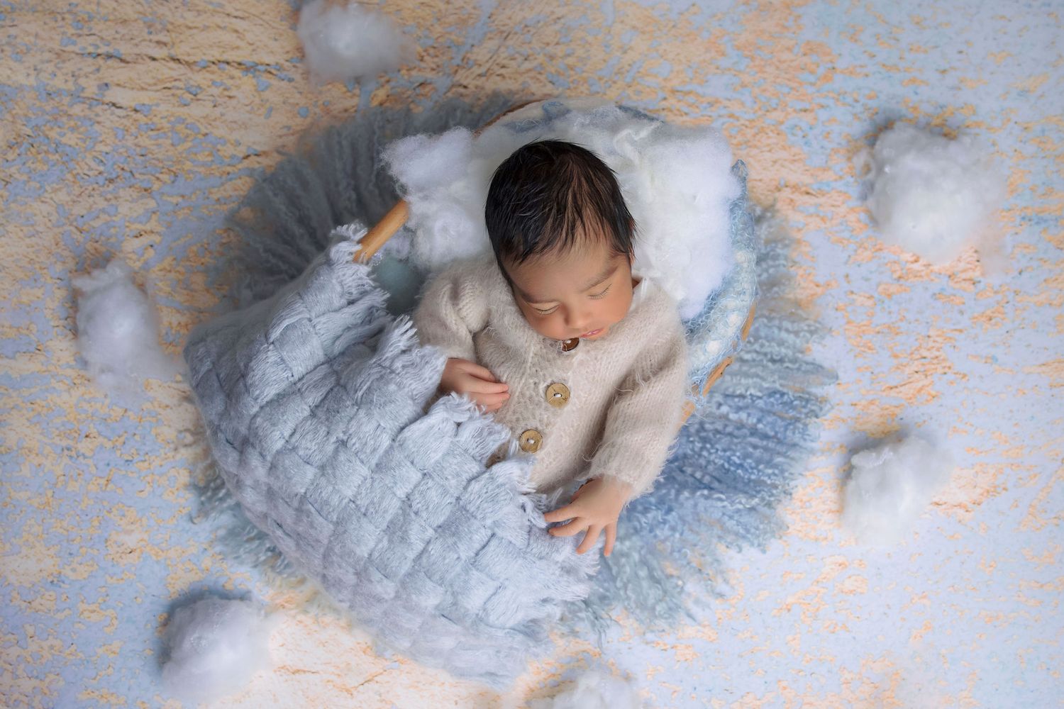 Sleeping newborn on blue textured blanket surrounded by cotton-like clouds.