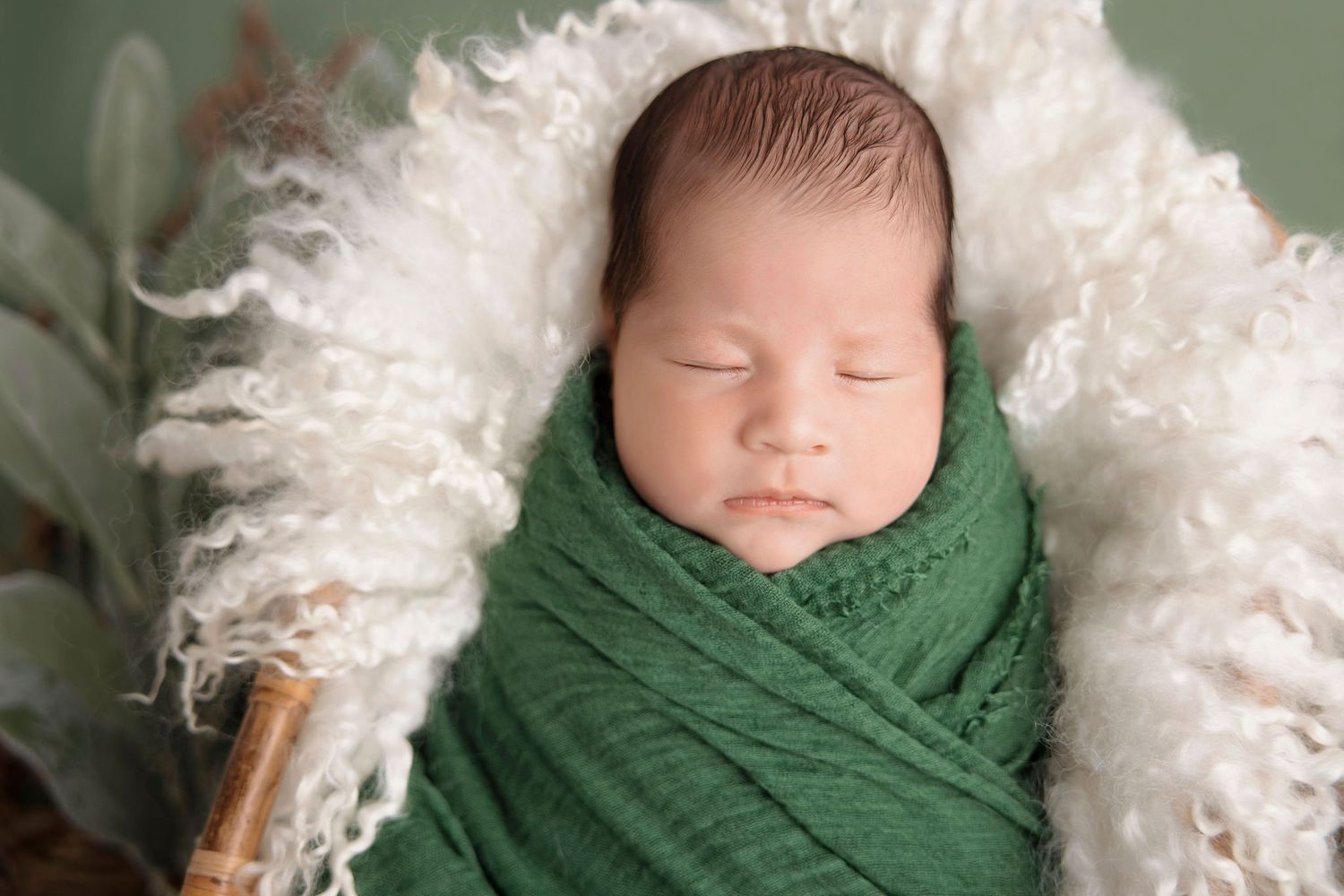 Newborn swaddled in green wrap resting on fluffy white fur backdrop.