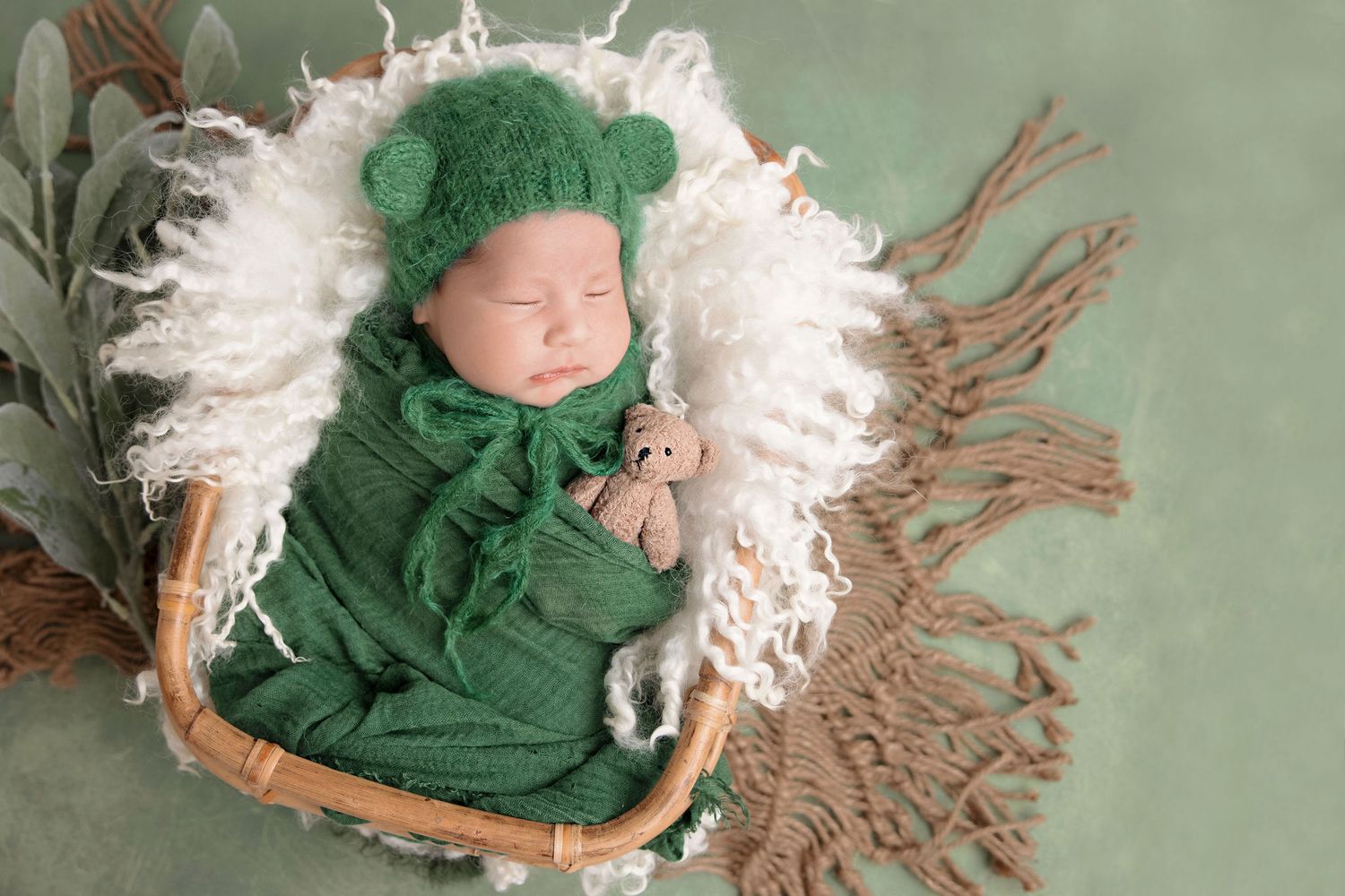Sleeping newborn in green knit outfit with bear ears on white fluffy blanket.