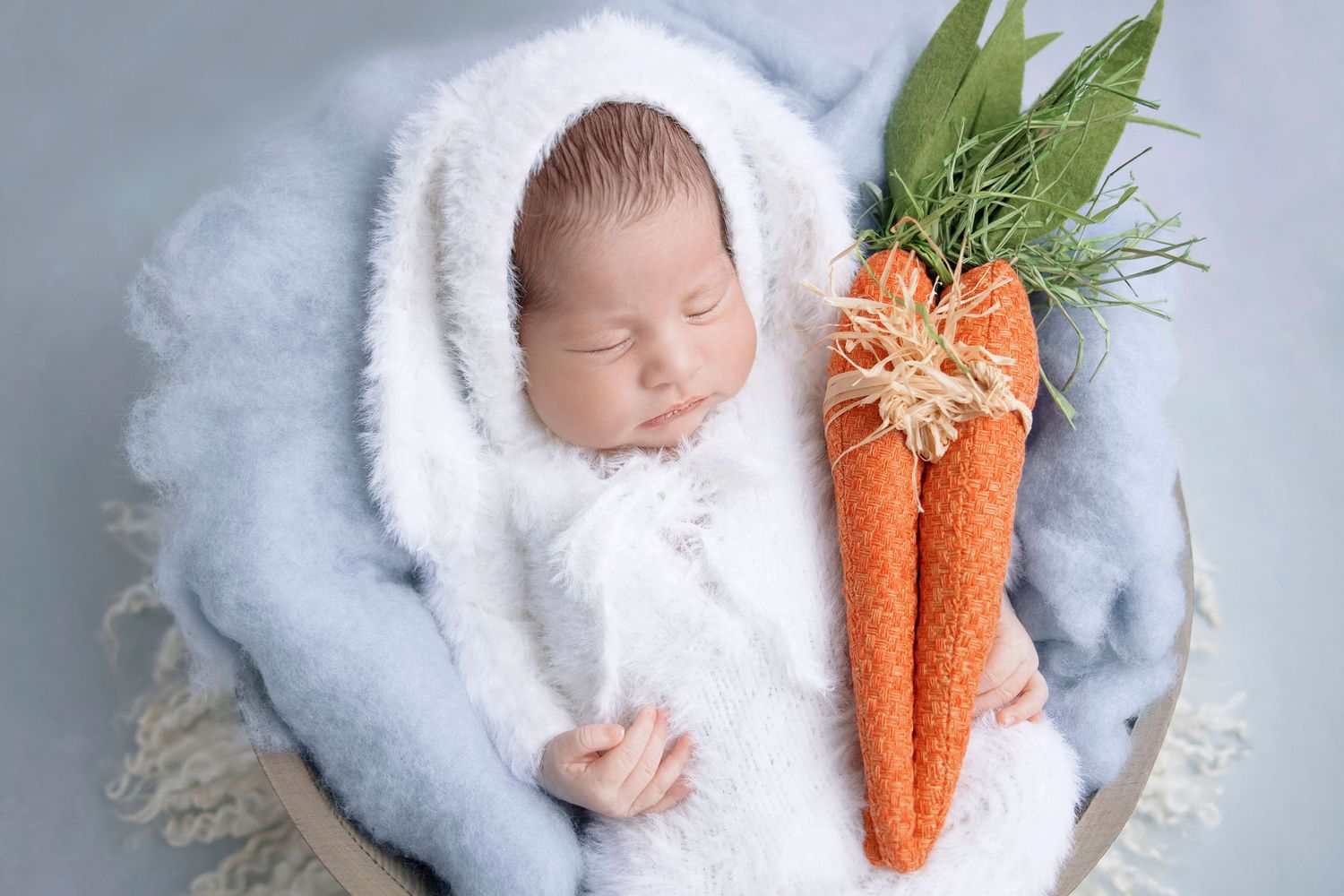 Sleeping newborn in white bunny outfit with decorative carrots on blue background.