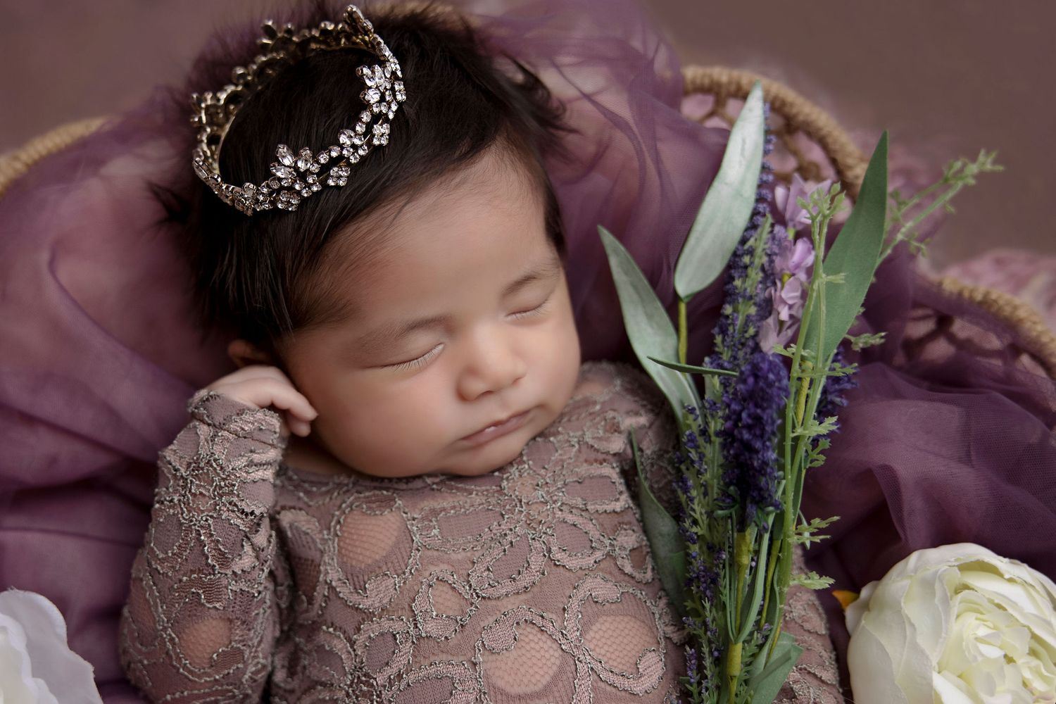 Sleeping newborn in lace dress and crown posed with purple flowers against mauve backdrop.