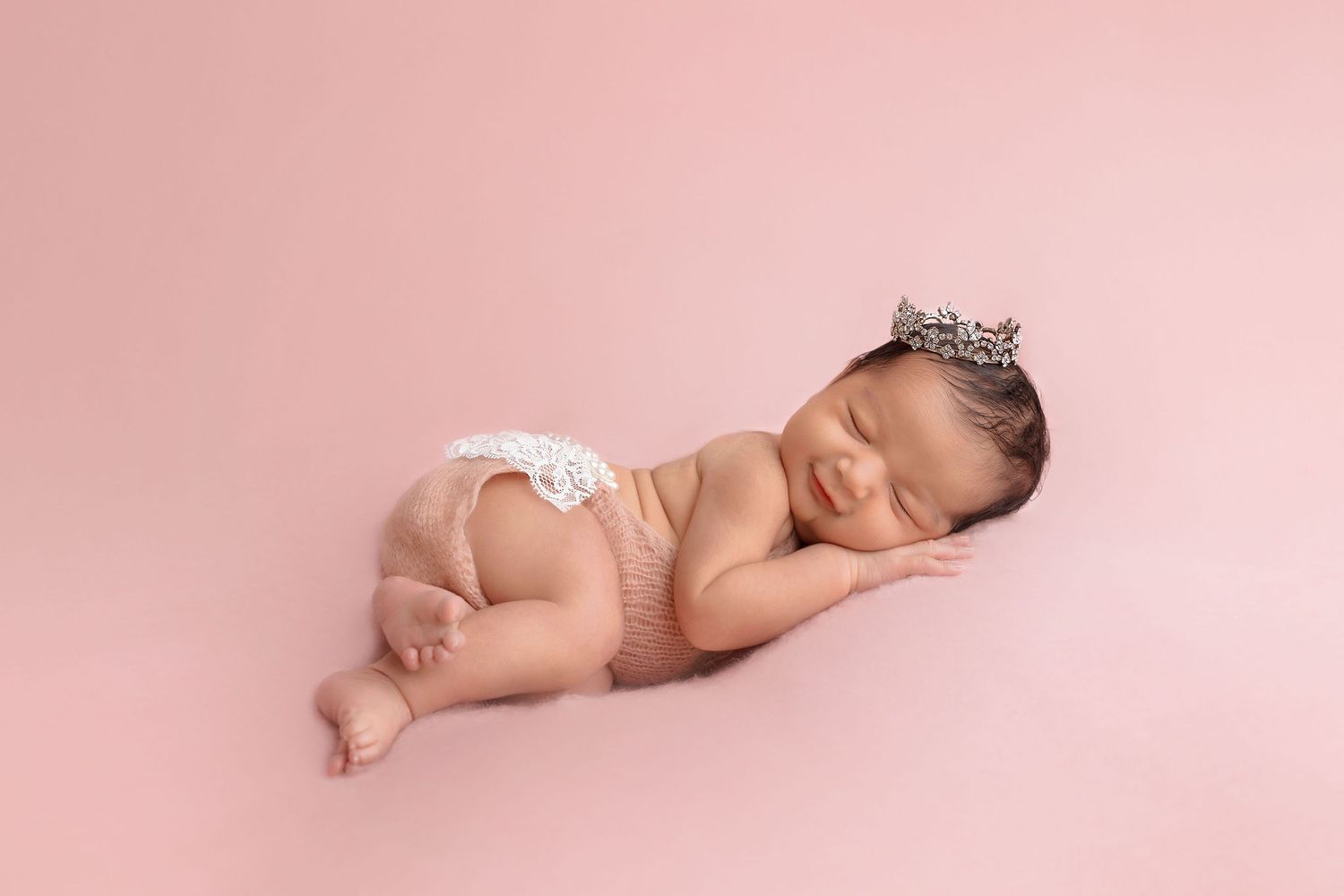Sleeping newborn wearing crown and diaper on pink backdrop.