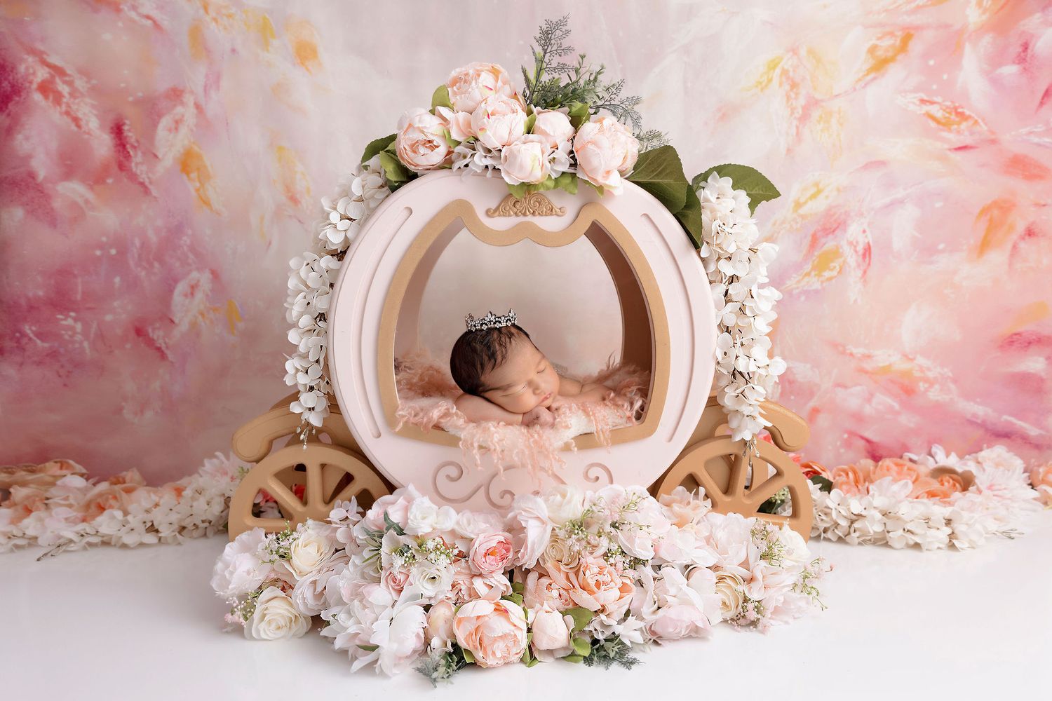 Newborn sleeping in ornate carriage prop decorated with pink roses against watercolor backdrop.