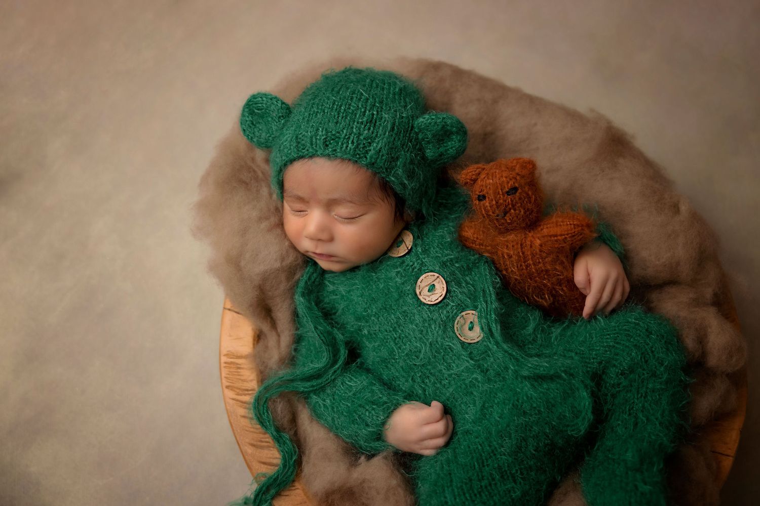 Sleeping baby in green knit bear outfit with teddy bear on brown blanket.