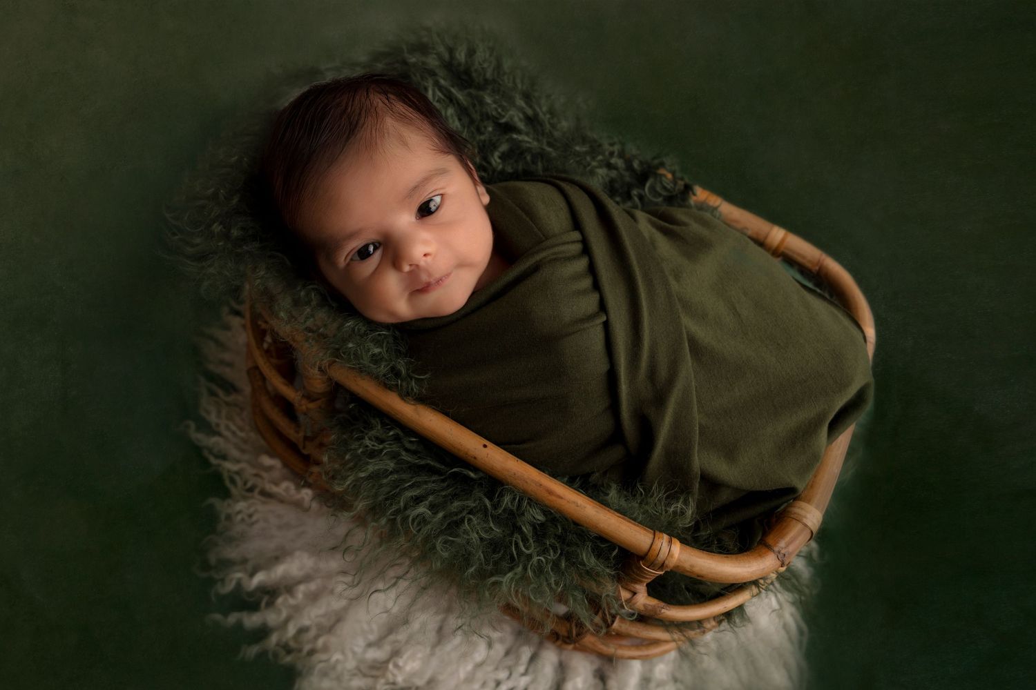 Baby resting in wooden basket with green backdrop and textured blanket.