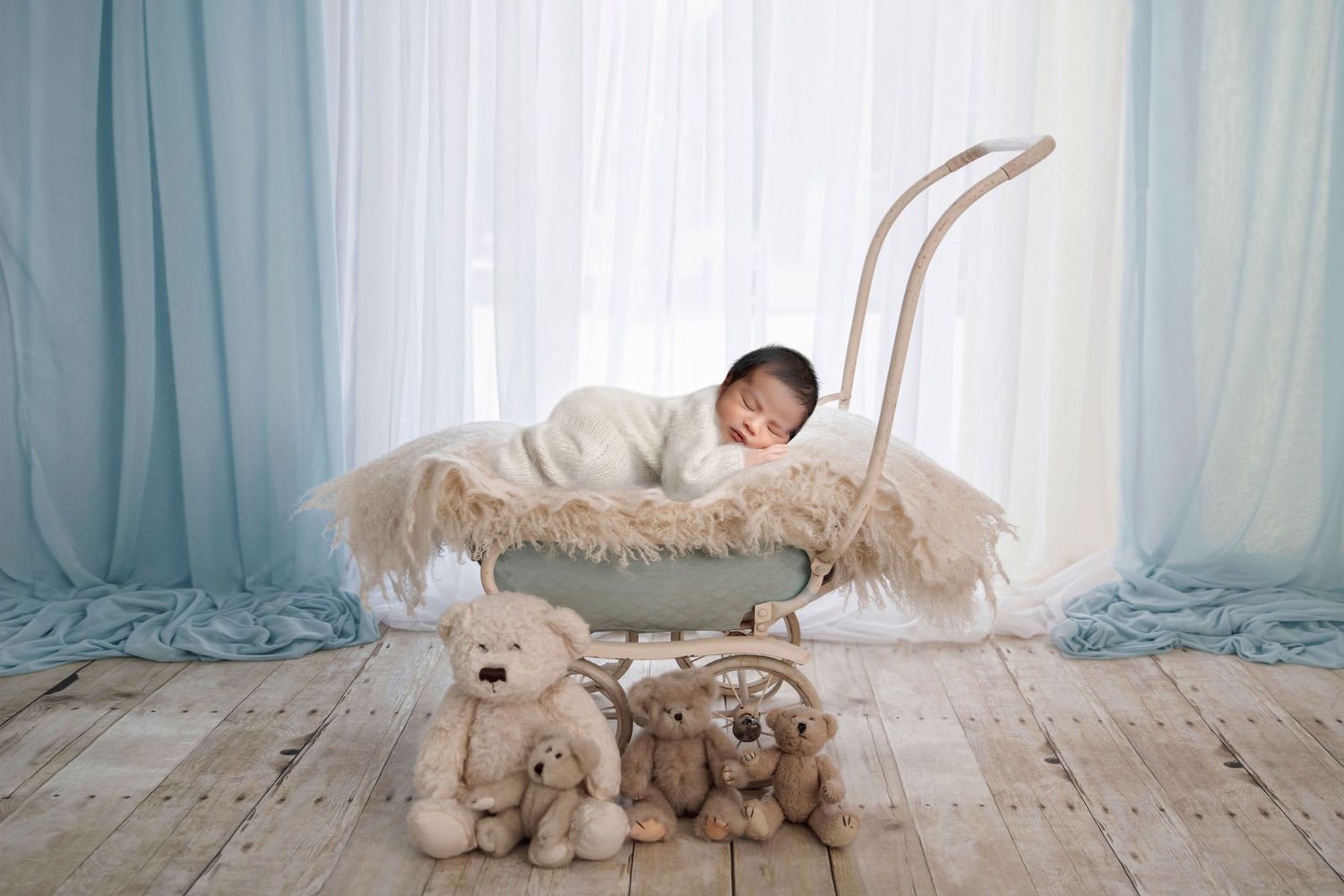 Baby resting in vintage pram with plush toys on wooden floor against blue curtain backdrop.