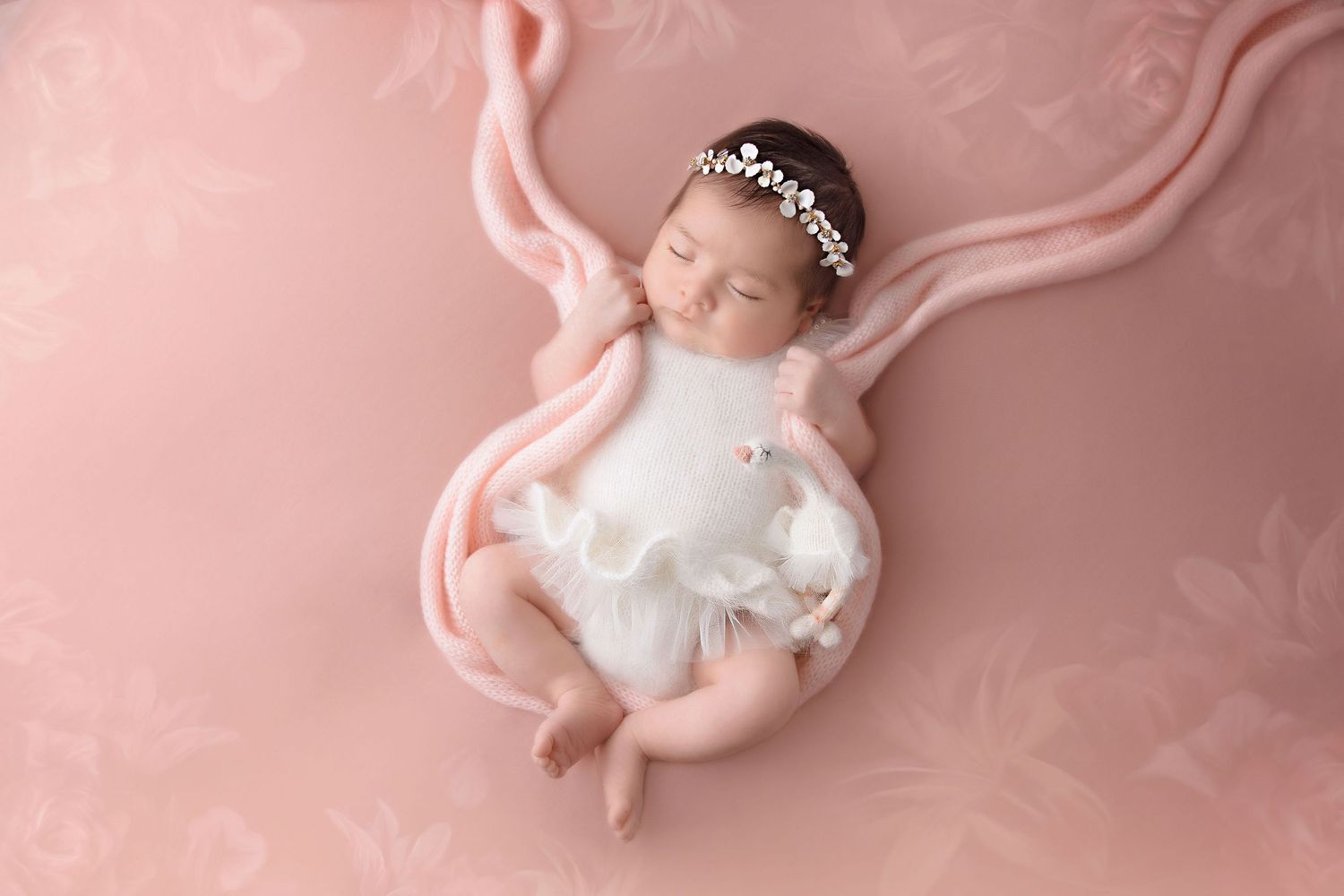 Sleeping newborn in white dress with delicate crown on pink backdrop with flowing fabric.
