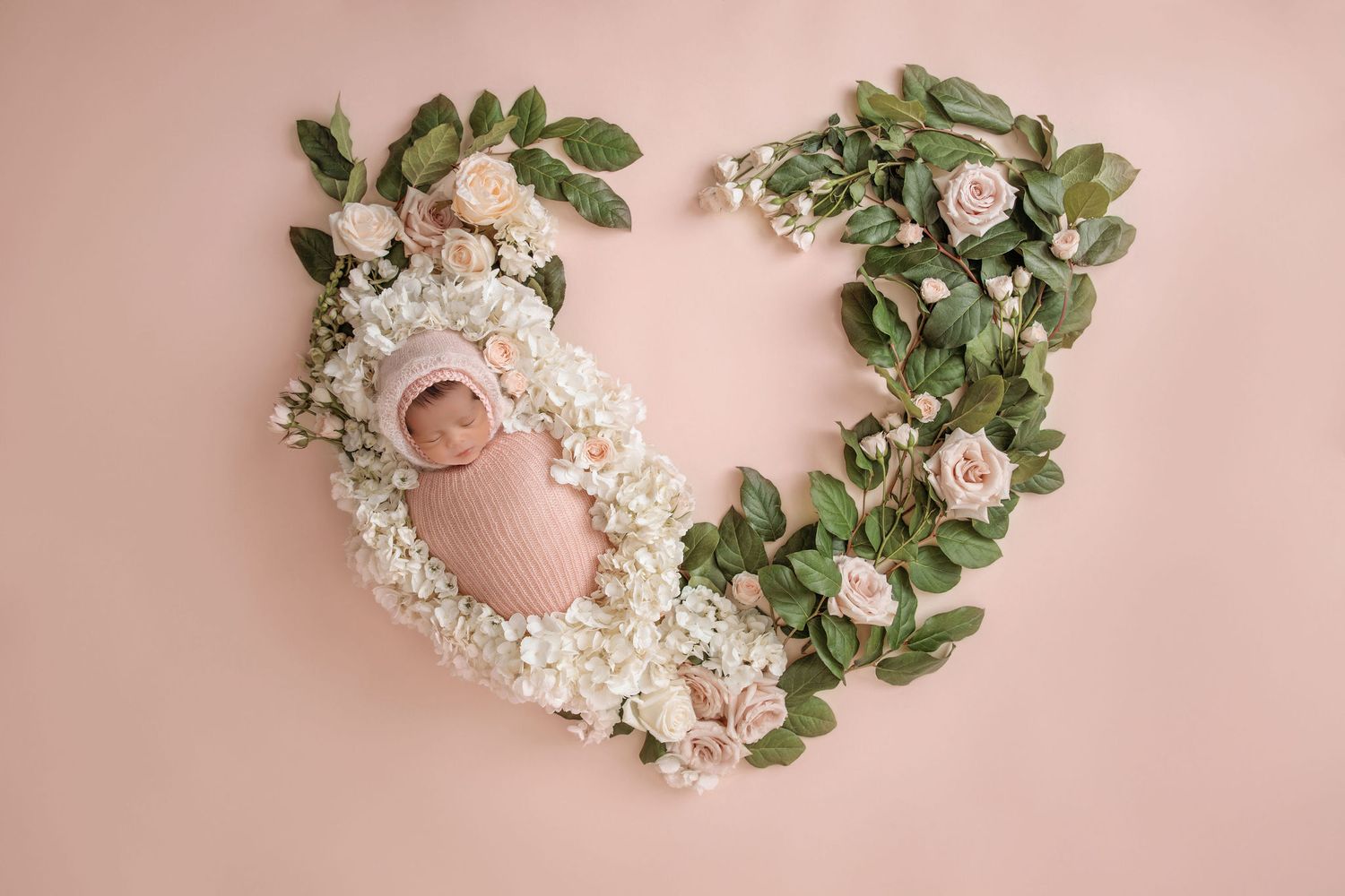 Newborn posed in floral wreath with greenery and white flowers on pink background.