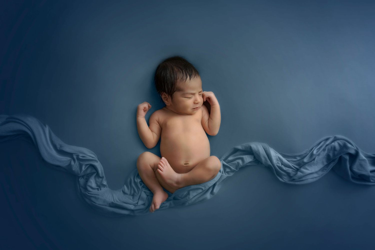 Newborn baby sleeping on blue fabric with artistic flowing drapery arrangement.