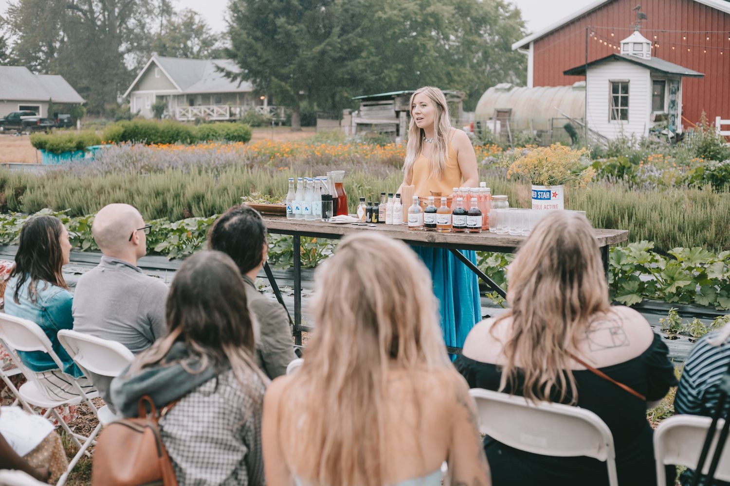 A woman in front of lavender and a barn teaching a bar class outside audience is in chairs facing her