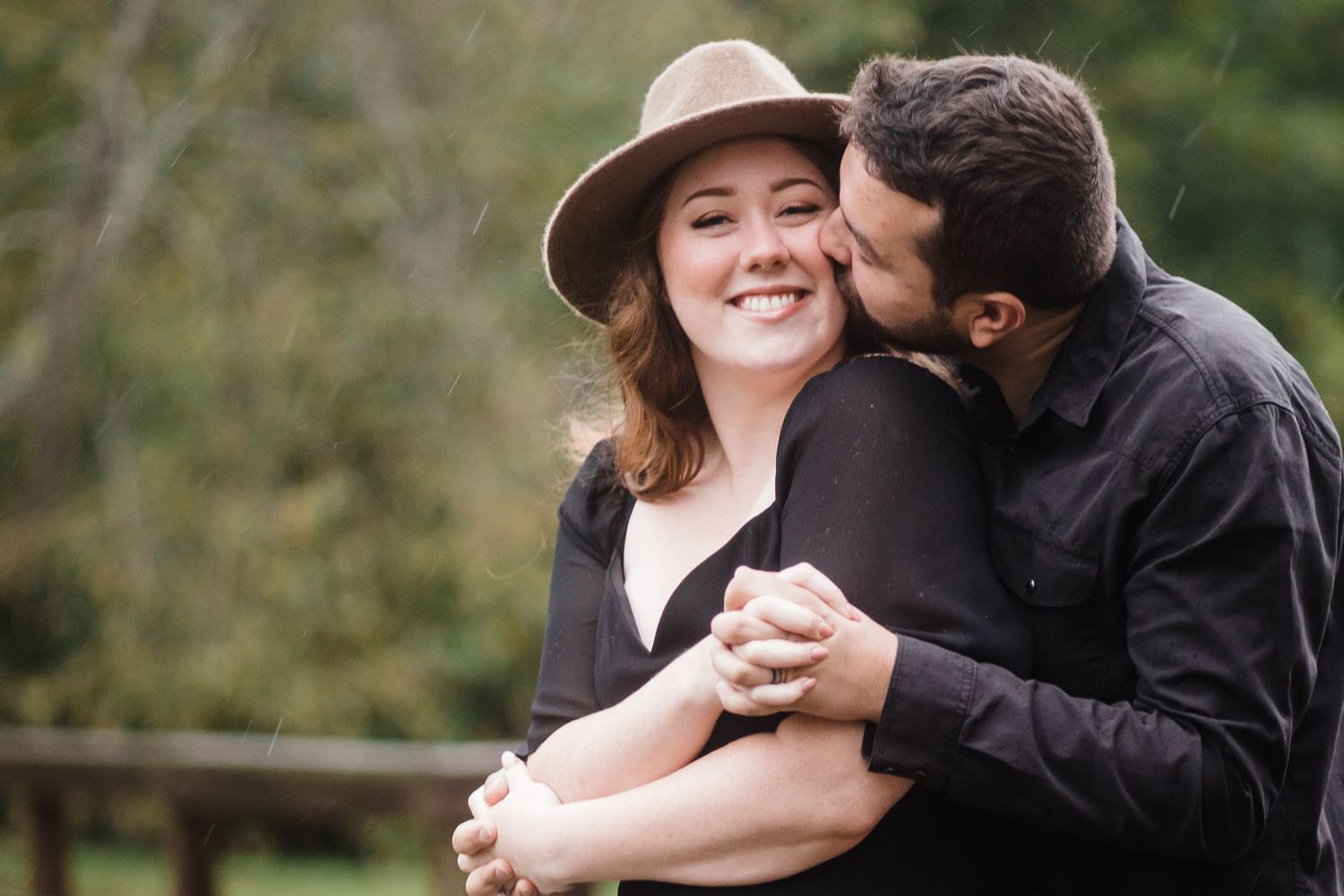 Couple in black clothing sharing intimate moment outdoors with person wearing brown hat.