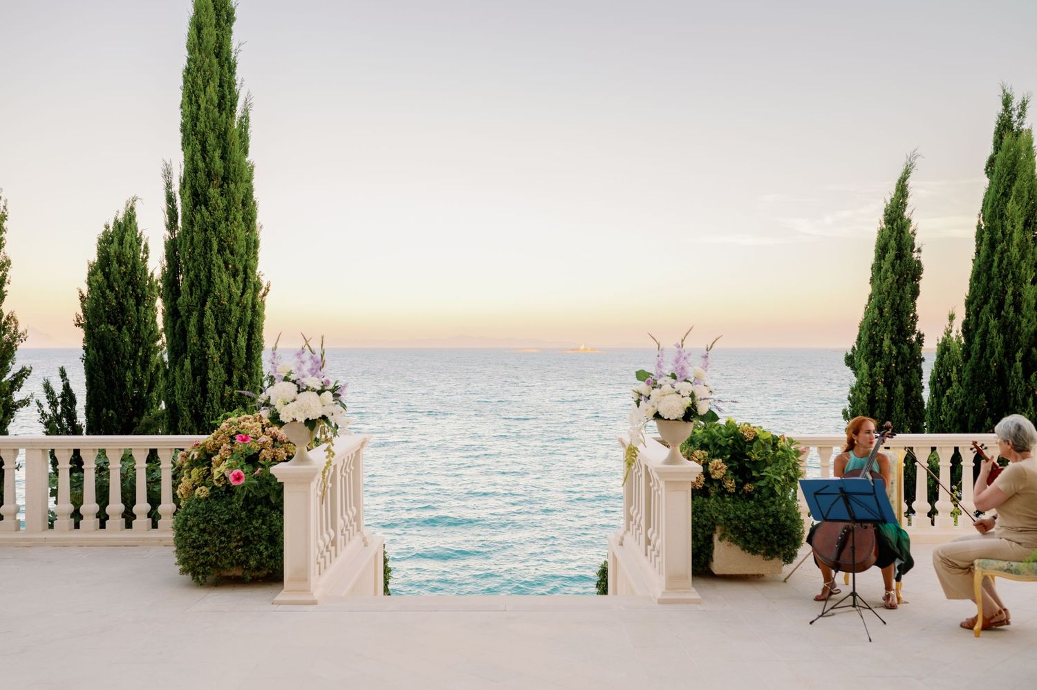 Mediterranean wedding ceremony setup with white columns and cypress trees overlooking the ocean at sunset.