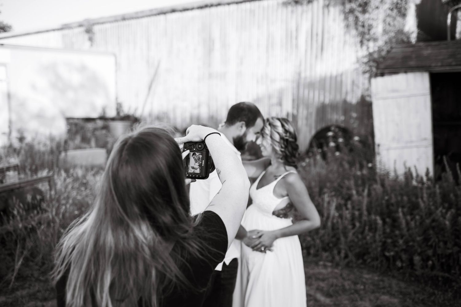 Black and white photo of a photographer capturing a couple embracing near a rustic barn outdoors.
