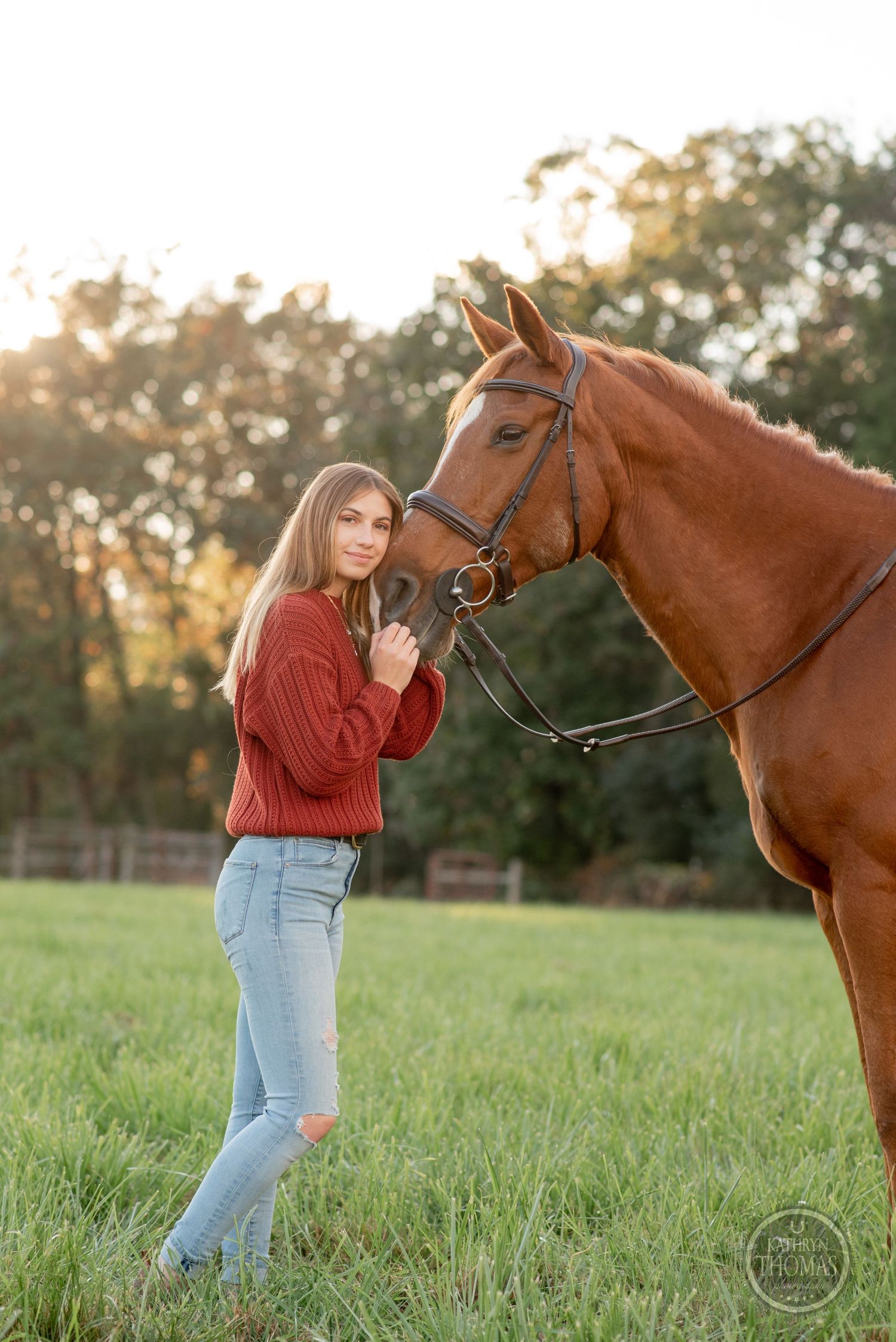 Chestnut horse with bridle stands close to rider in red sweater during sunset photoshoot.