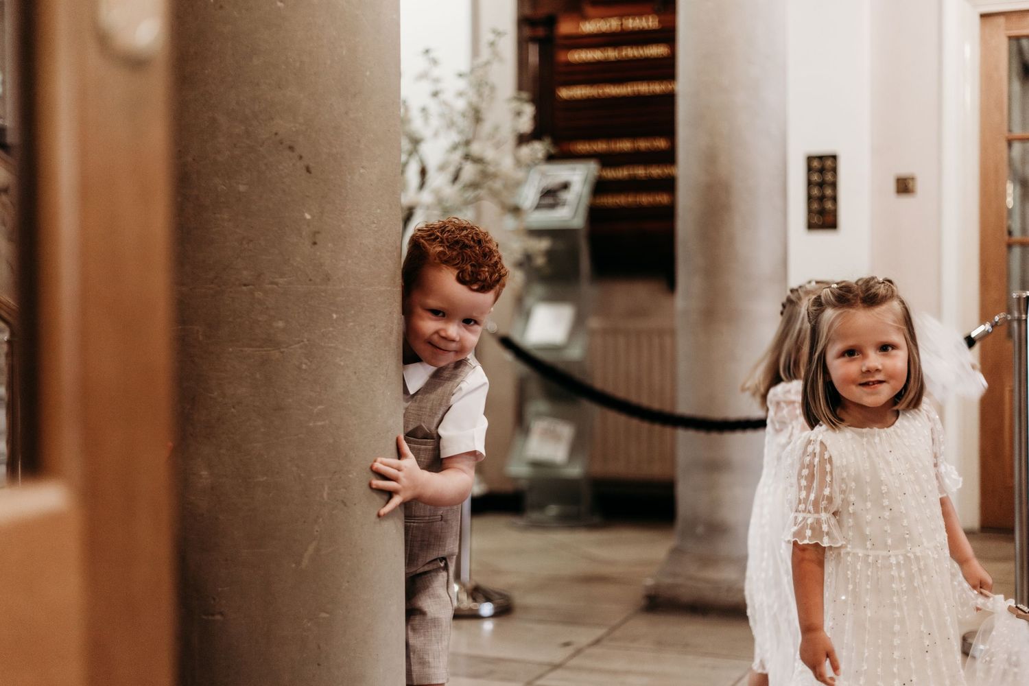 Two children in formal attire playfully peek around a classical column.