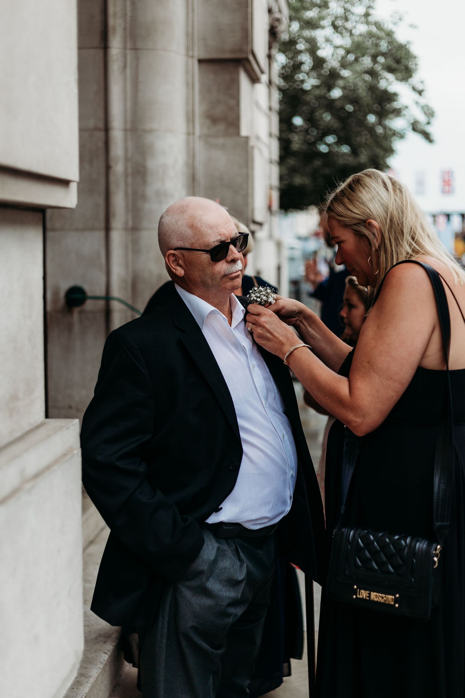 Someone adjusts a boutonniere on a person wearing sunglasses and a dark suit outdoors.