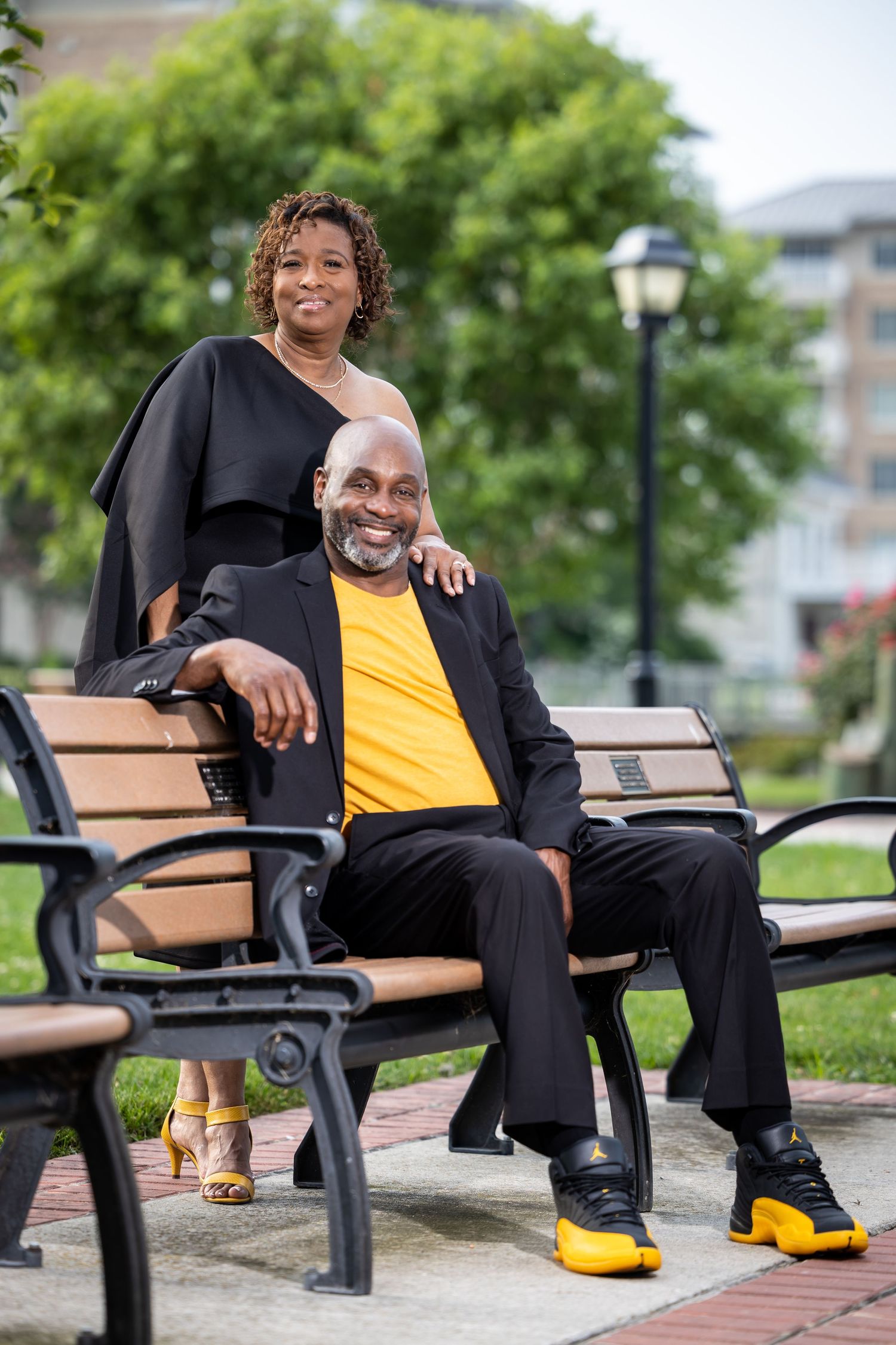 A couple in black attire poses playfully on a park bench with yellow accents in their outfits.